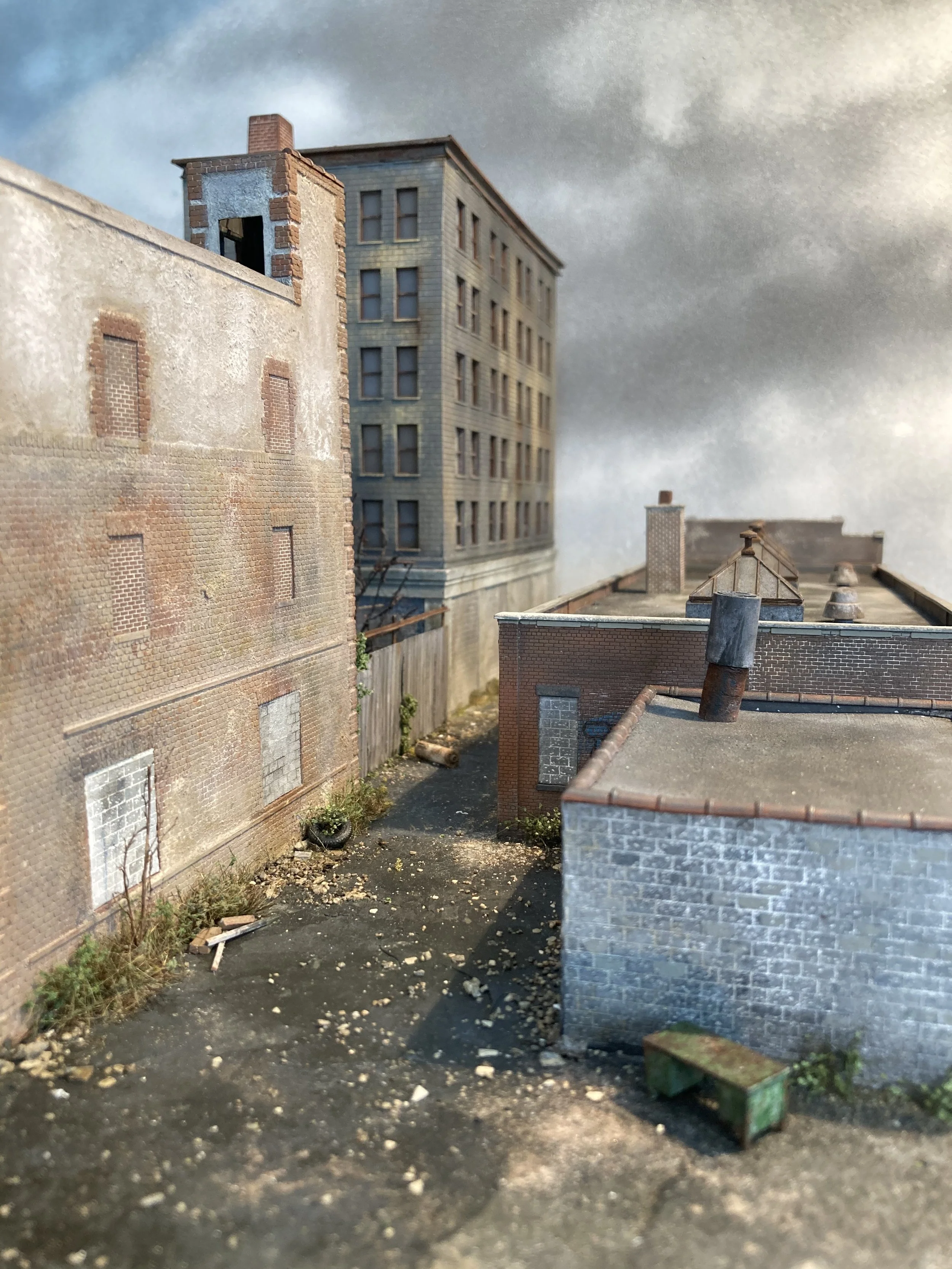 A miniature model of an alleyway with buildings, a brick wall, and a small green box on the ground.