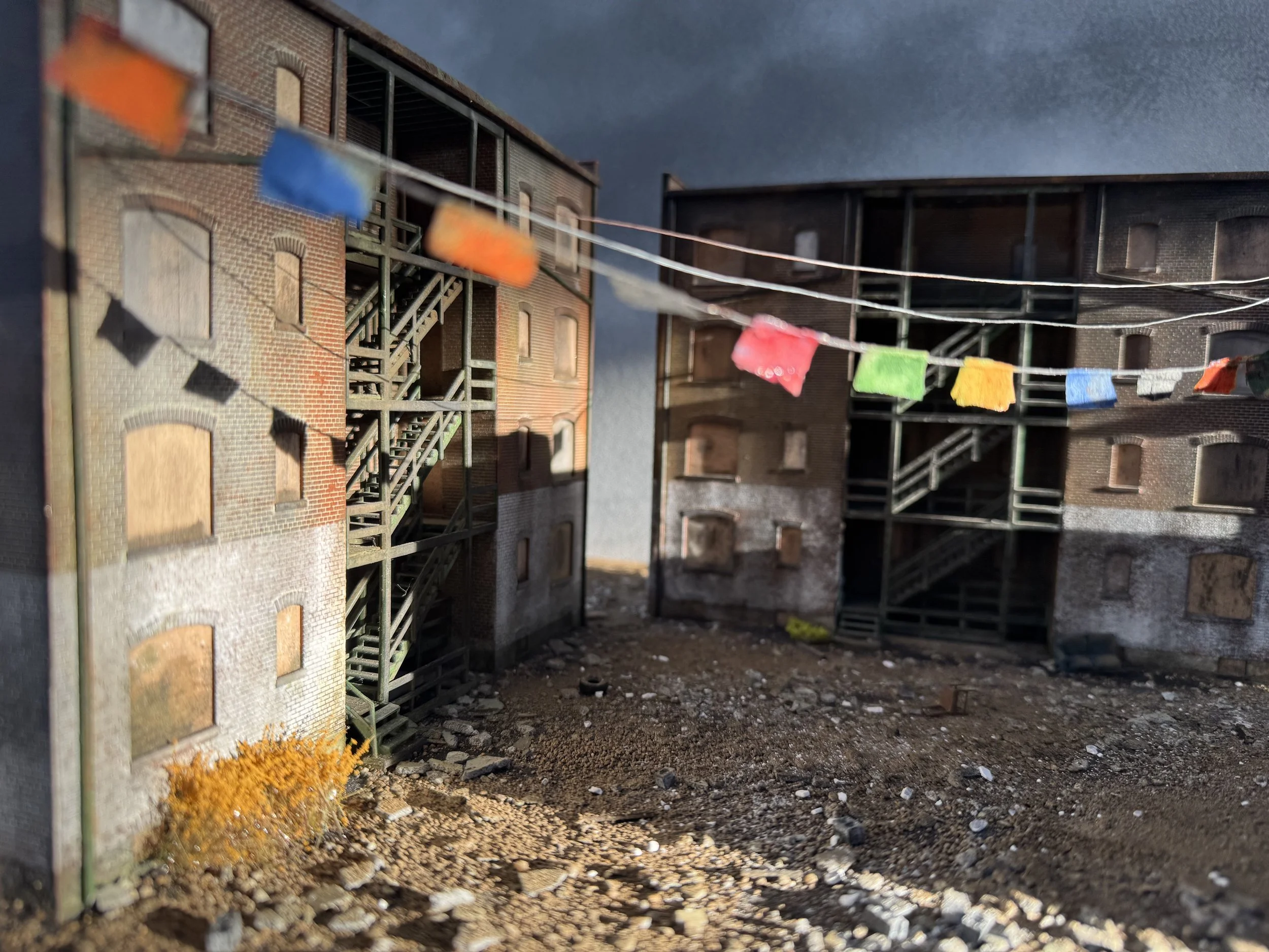 Model urban apartment buildings with fire escapes, colorful flags hung across. Ground features dirt and scattered small rocks, with a small bush at the lower left corner. Overcast sky.