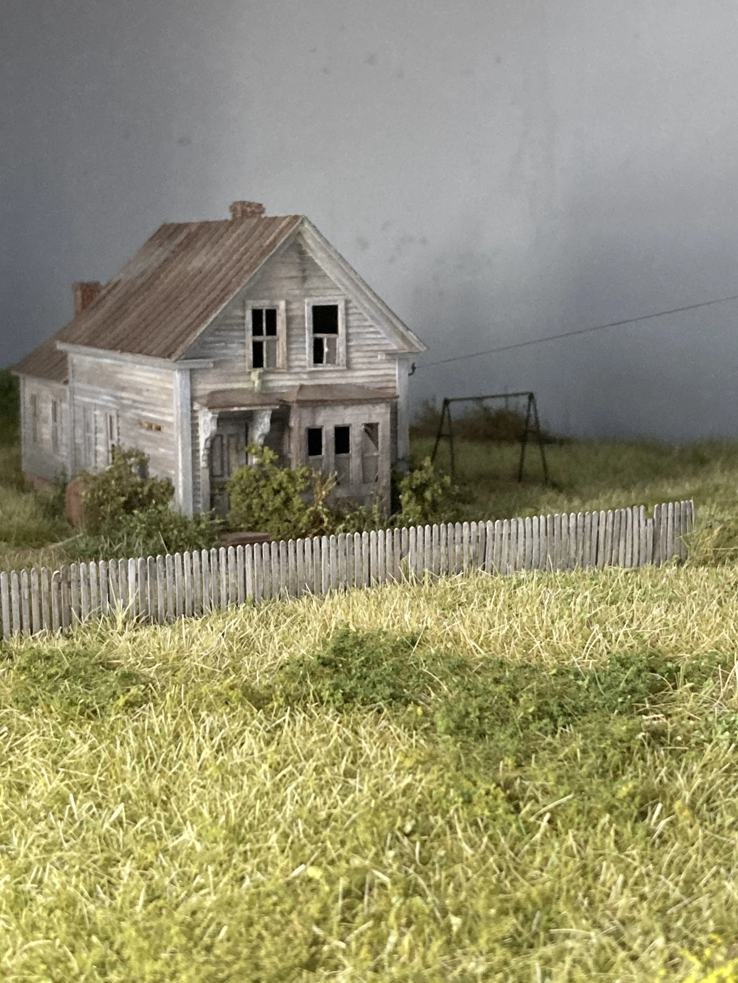 A weathered, abandoned wooden house with broken windows, a rusted roof, surrounded by overgrown grass and a white picket fence, under a dark, stormy sky.