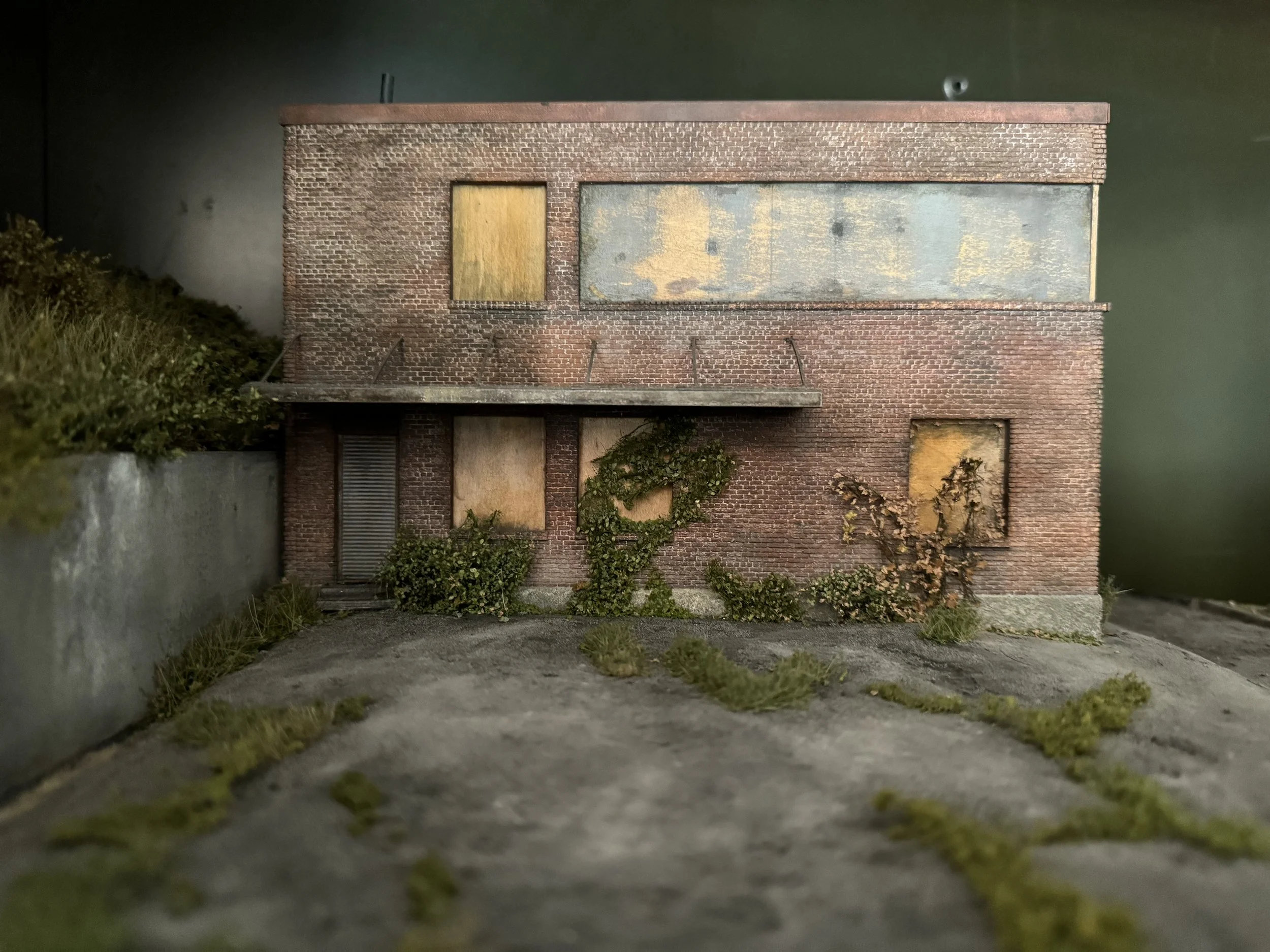 A model of an abandoned brick building with boarded windows and some greenery growing at its base, seen from a low angle close to the ground.
