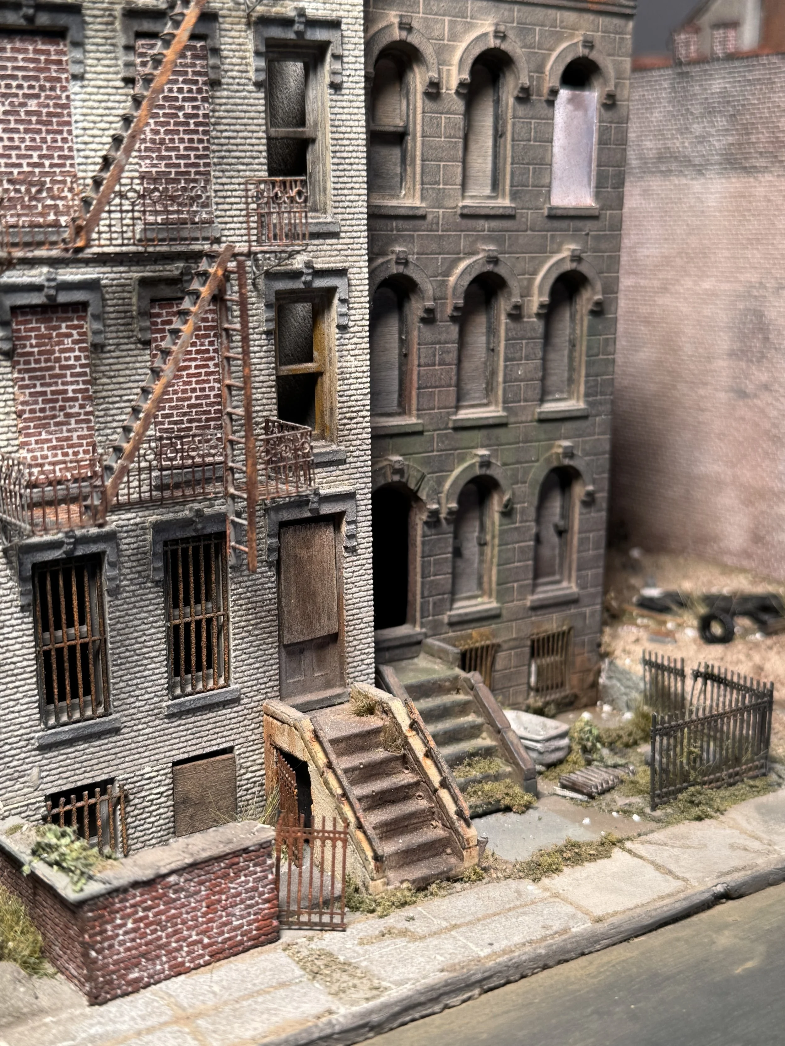 A detailed miniature model of a row of old urban buildings with brick exteriors, fire escape, and stairs leading to small doors.