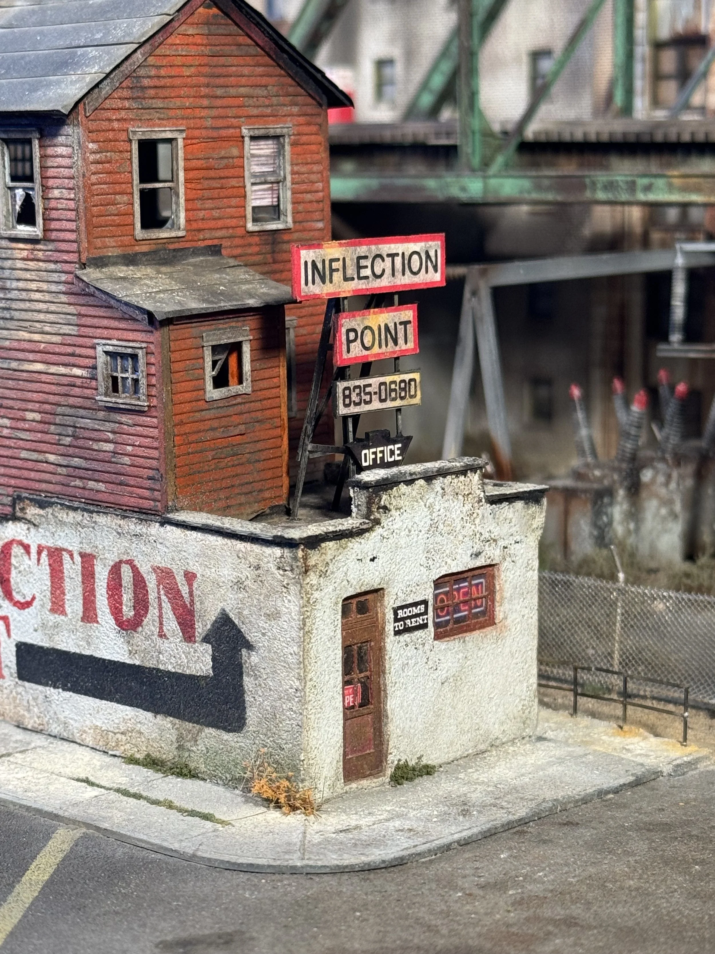 A miniature model of a two-story brick building with a sign that reads 'INFECTION POINT' and a phone number, next to a sidewalk and a street.