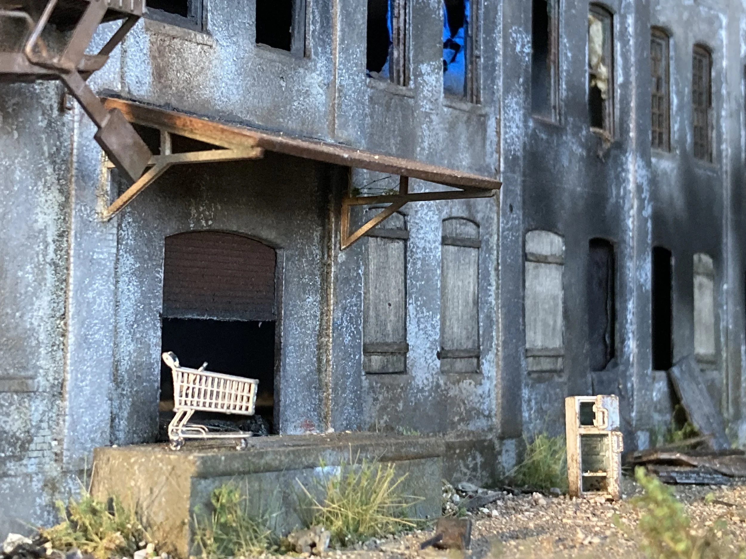 The image depicts a weathered, abandoned building with a stone foundation and a painted, peeling exterior. Several windows are visible, some broken or missing glass. There are wooden and metal objects scattered on the ground, including chairs and pla