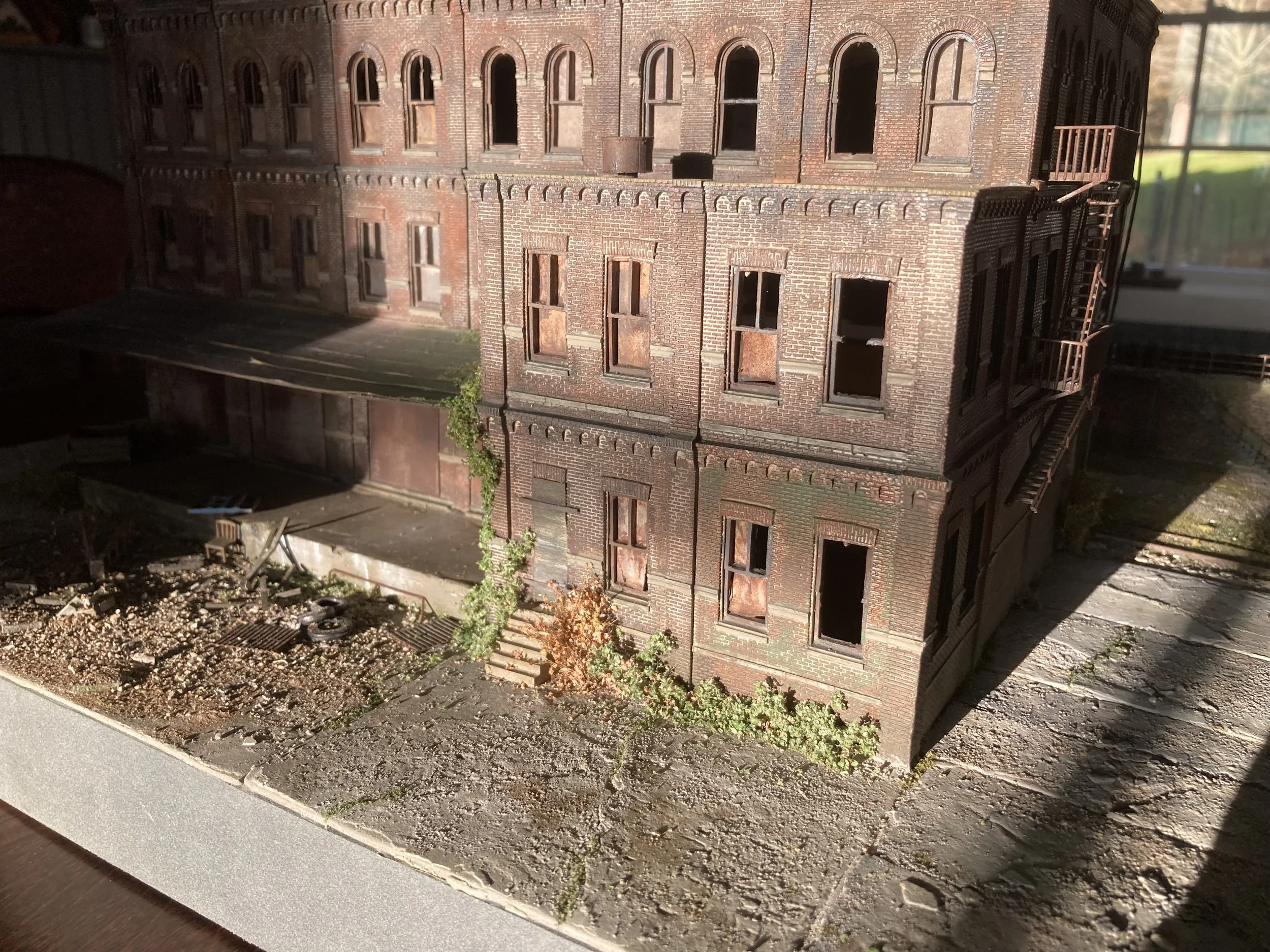 A detailed model of a red brick building, resembling an old apartment or commercial building, with multiple floors and numerous windows, some of which appear to have boarded-up or missing glass. The building features an exterior fire escape ladder on