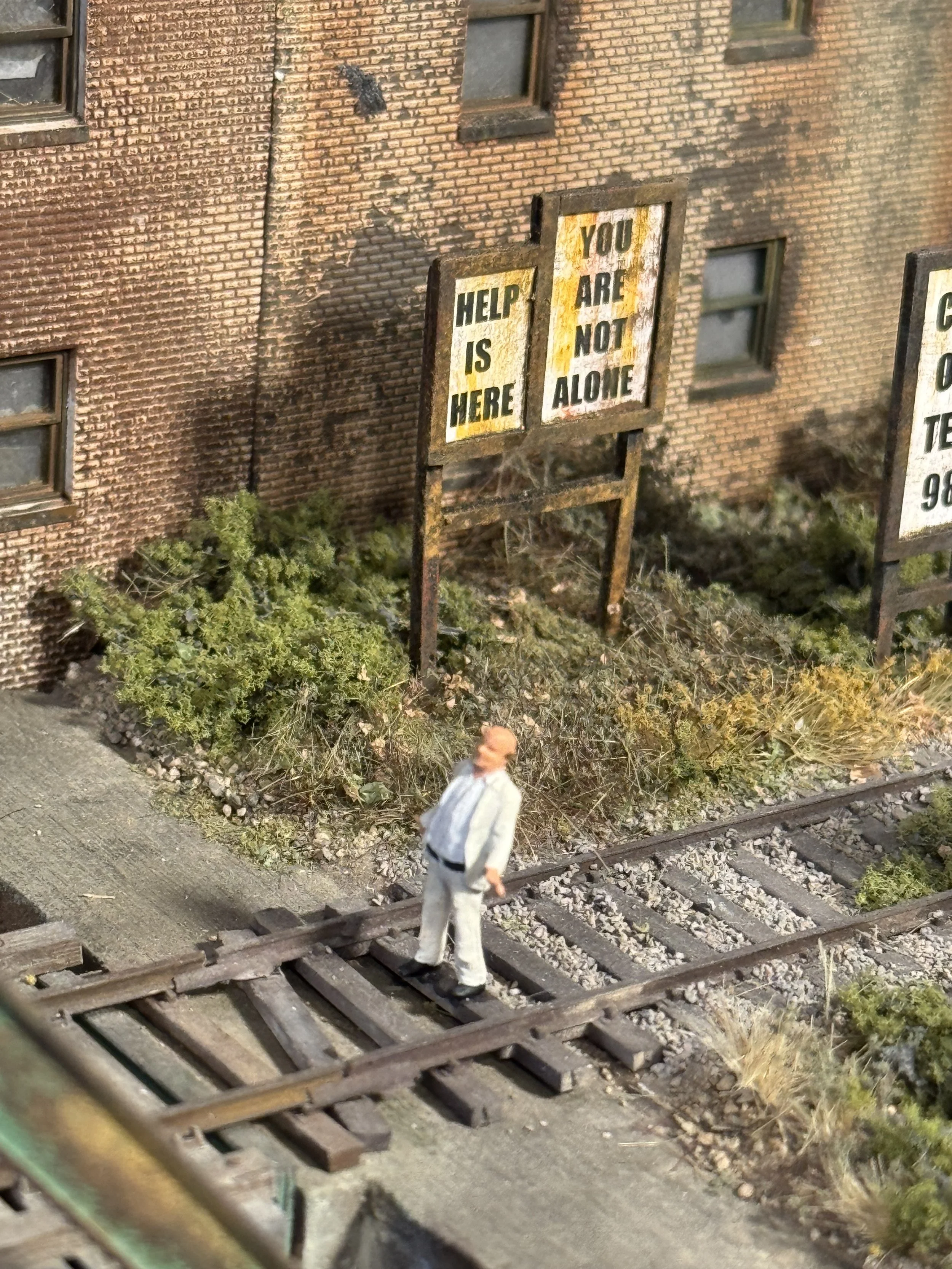 Miniature scene of a man in a gray suit walking on a railway track beside a brick building with graffiti and a weathered sign that says "Help is Here" and "You are not alone."