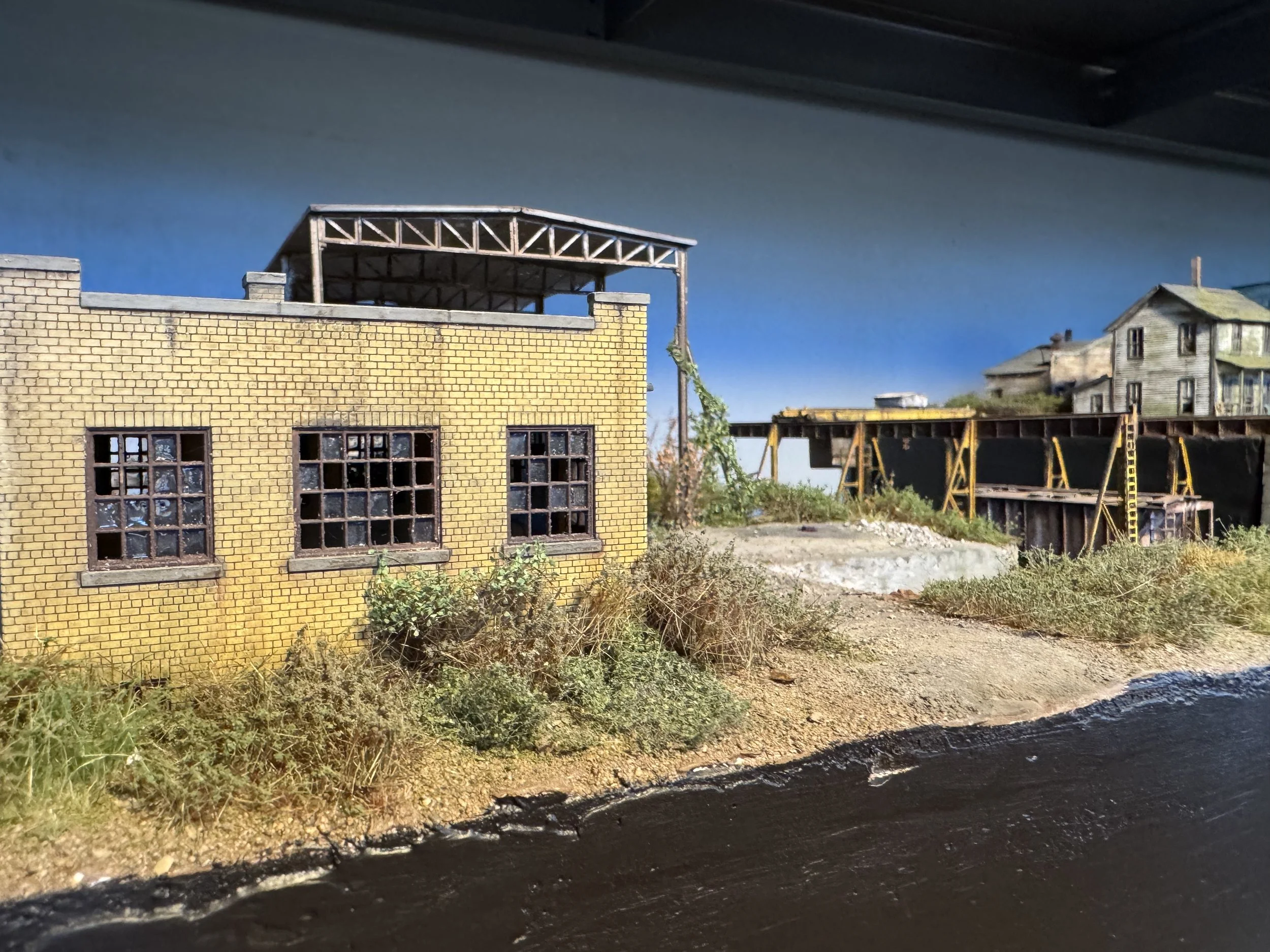 A miniature model of a yellow brick building with large windows next to a railway track with a train in the background under a cloudy sky.
