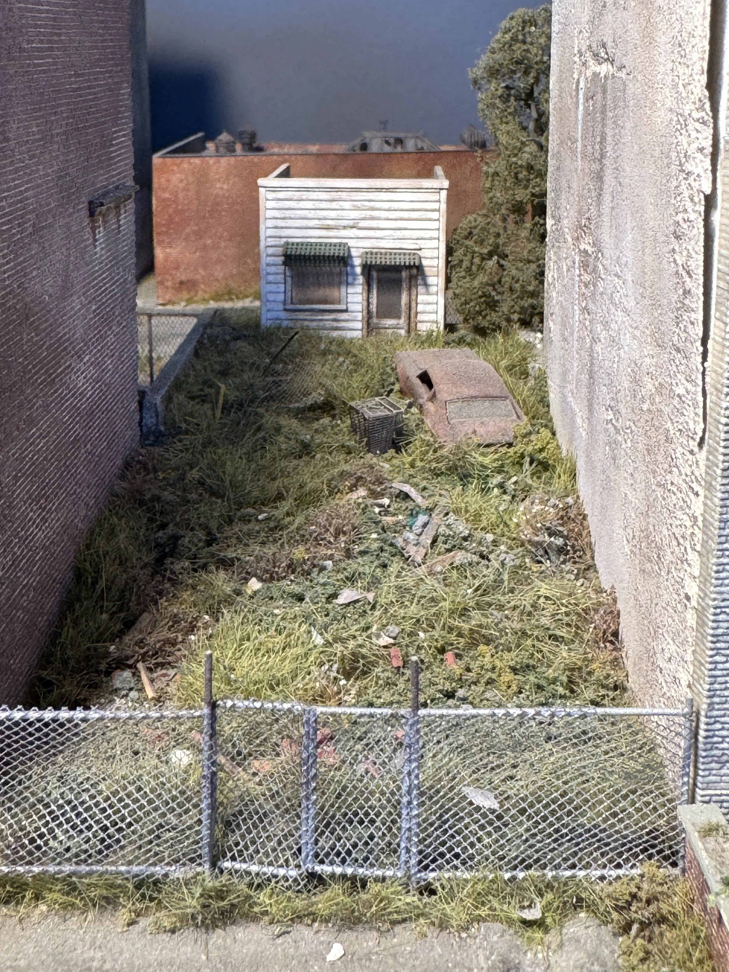 A small, white, weathered shed with green awnings over the windows and door, surrounded by overgrown grass and weeds, enclosed by a chain-link fence, with brick and concrete walls on either side and a building with a brick wall in the background.