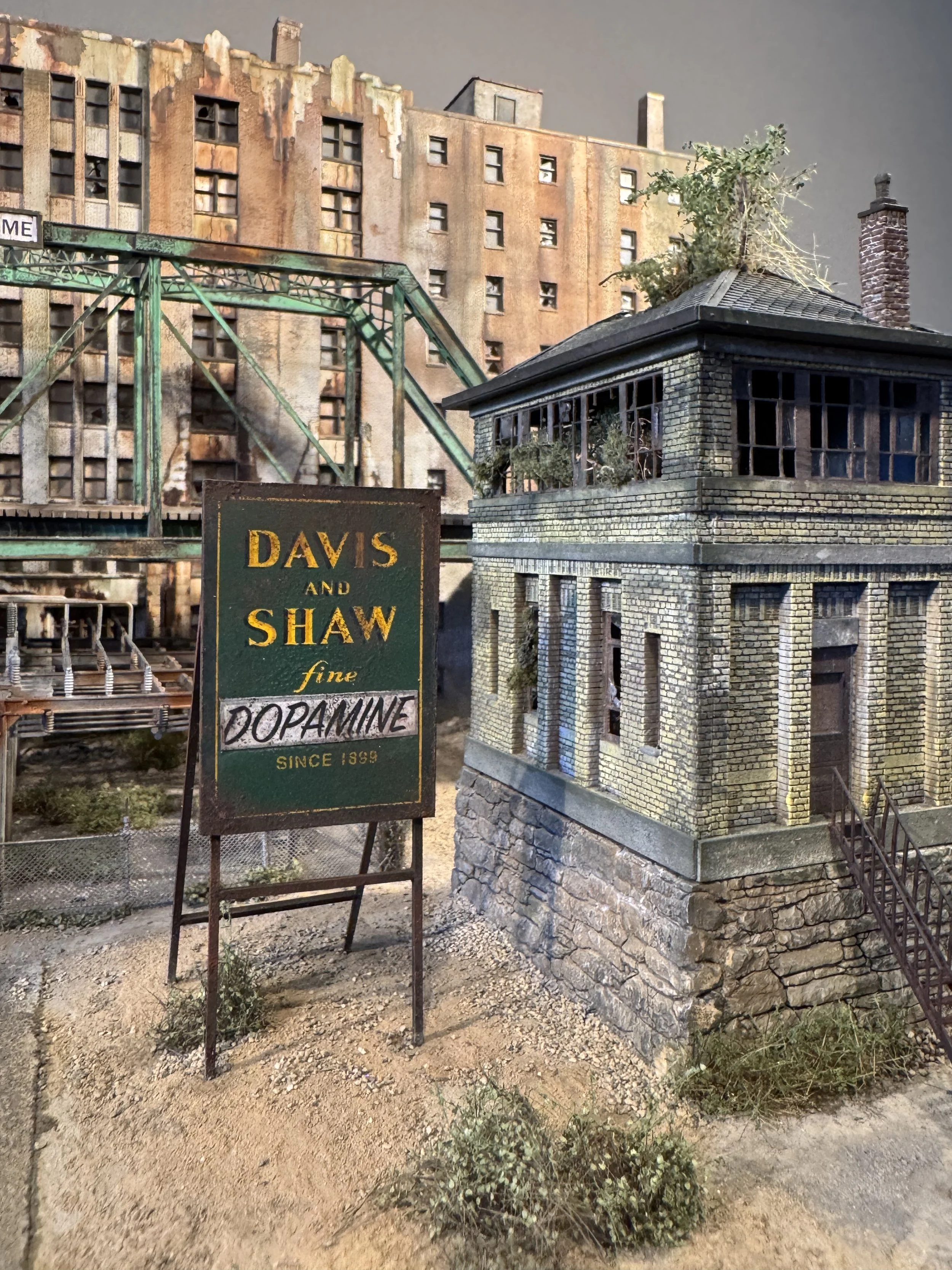 Miniature model of an old brick building with columns, adjacent to a smaller brick structure, in a setting with dirt ground and sparse bushes, with a sign for Davis and Shaw forensic dopamine.
