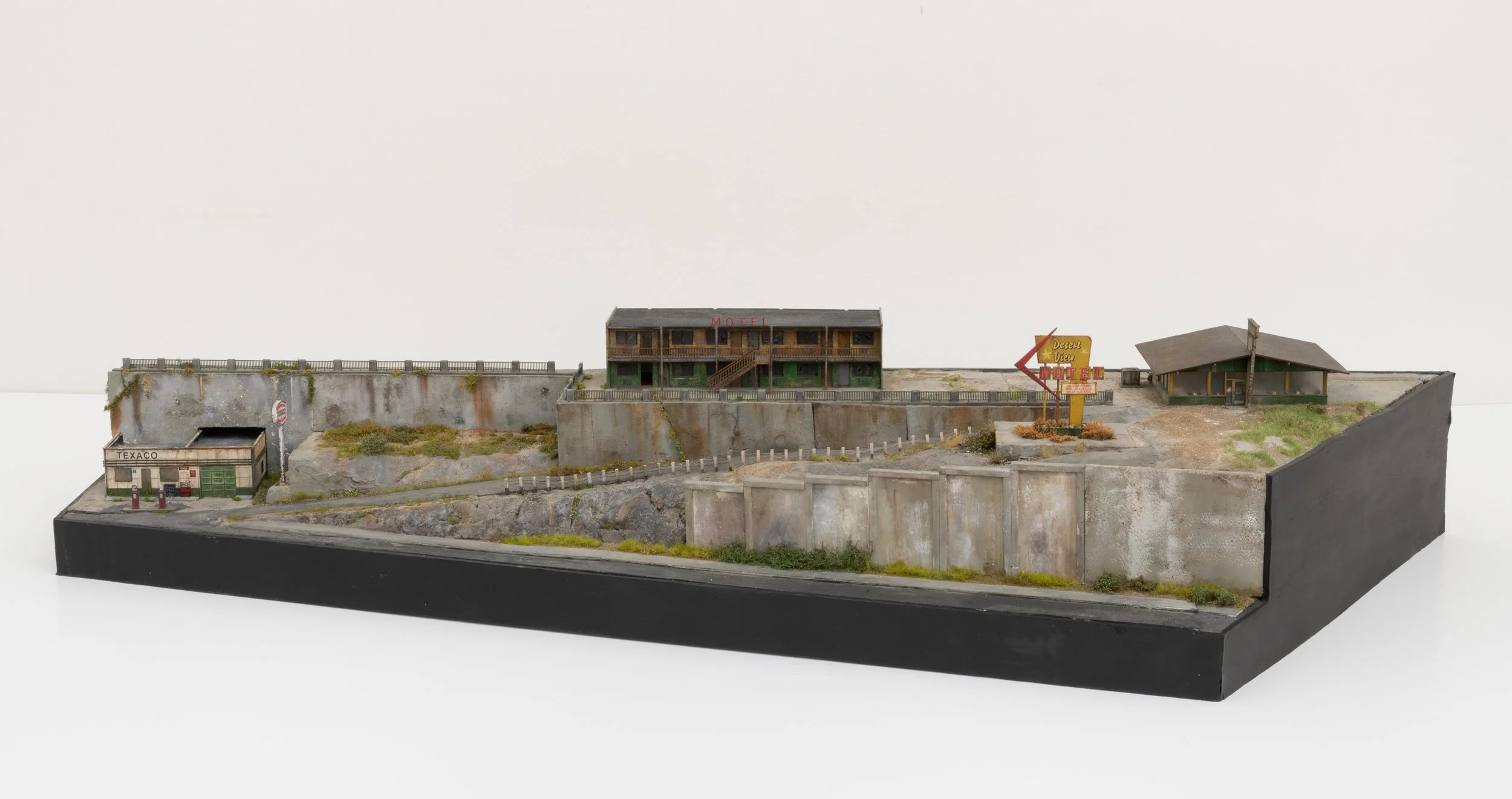 Miniature model of an old, run-down motel and gas station on a concrete base, with weathered buildings, overgrown plants, and signs indicating a desert view motel and Texaco gas station.