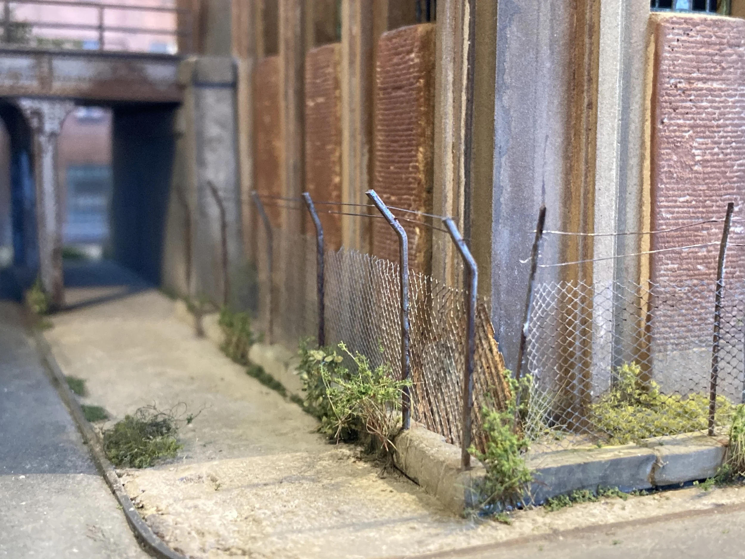 A chain-link fence with bent and damaged sections along a sidewalk, with small weeds and grass growing near the base of the fence, and a brick and concrete wall behind the fence.