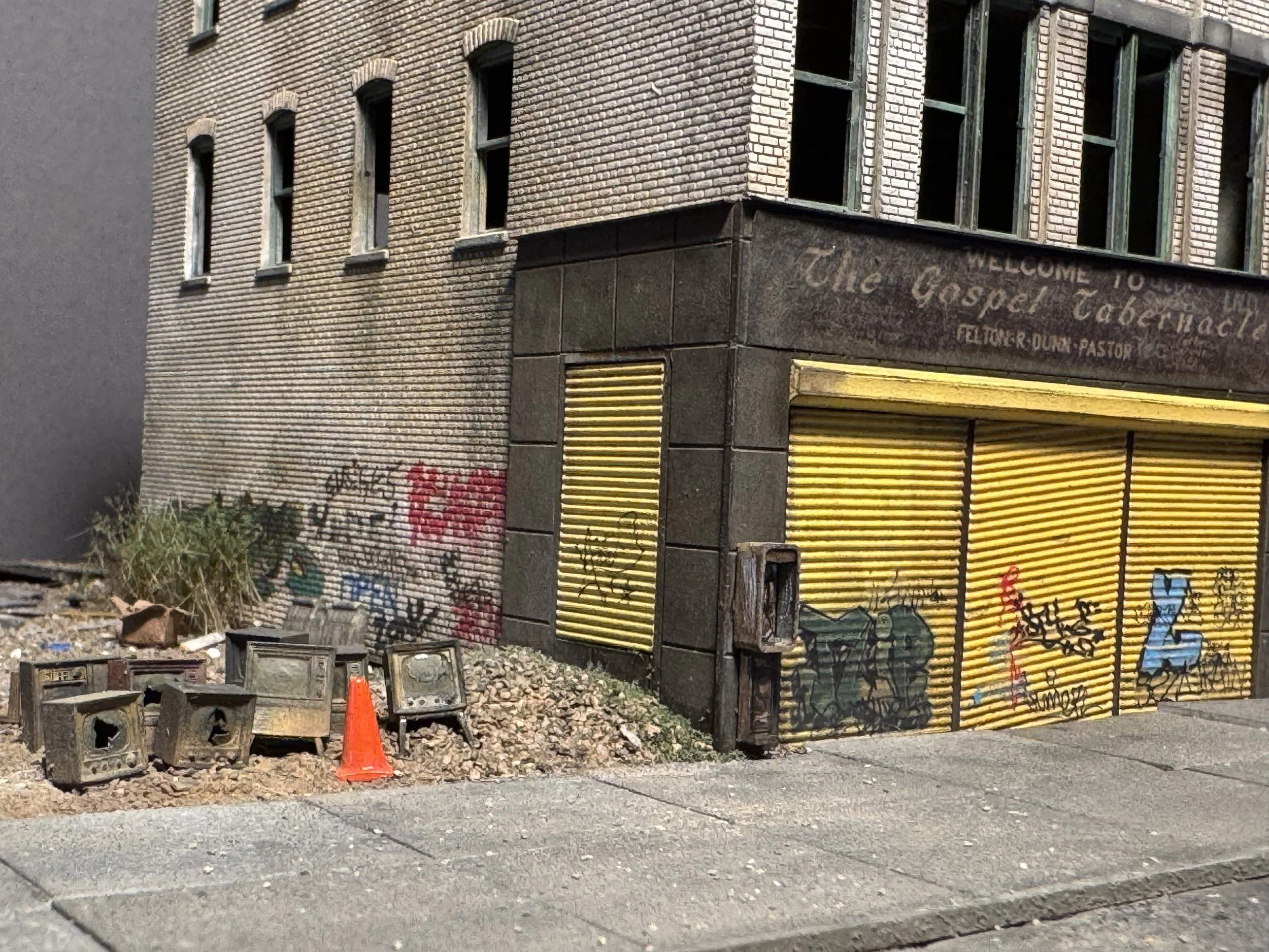 An abandoned building with boarded-up windows and graffiti on the wall, surrounded by construction debris, orange cones, and cinder blocks in the foreground.