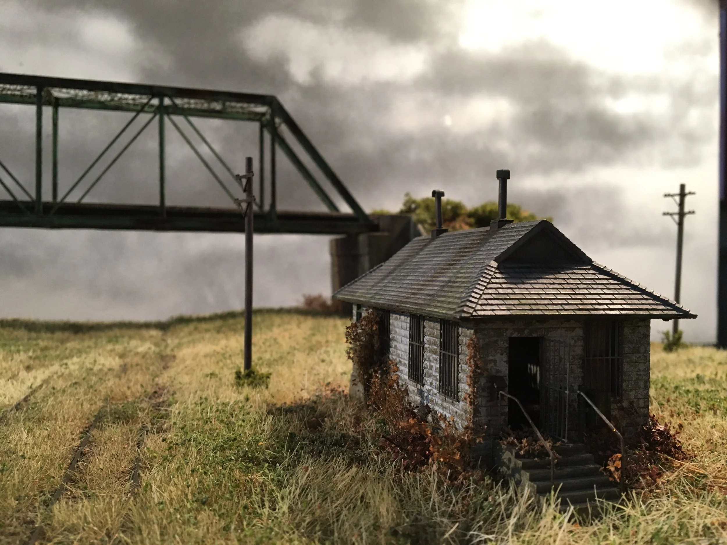 A model of a small brick house with a gray shingled roof, surrounded by grass, with two small chimneys on the roof. In the background, there is a large green metal bridge structure and utility poles against a cloudy sky.