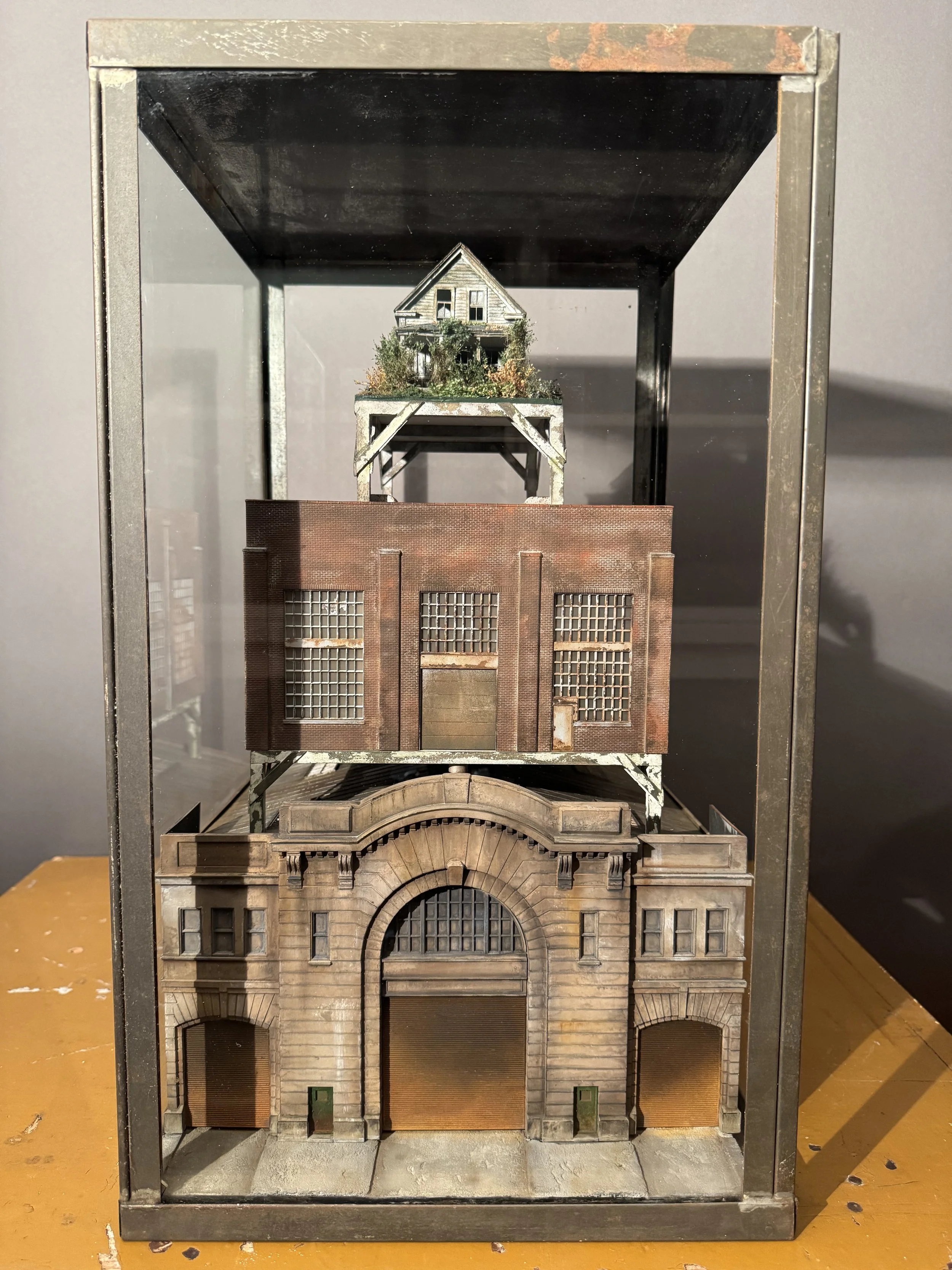 A miniature model of a multi-story city building with an arched entrance, brick and stone facade, and roller shutters, housed in a glass and metal display case.