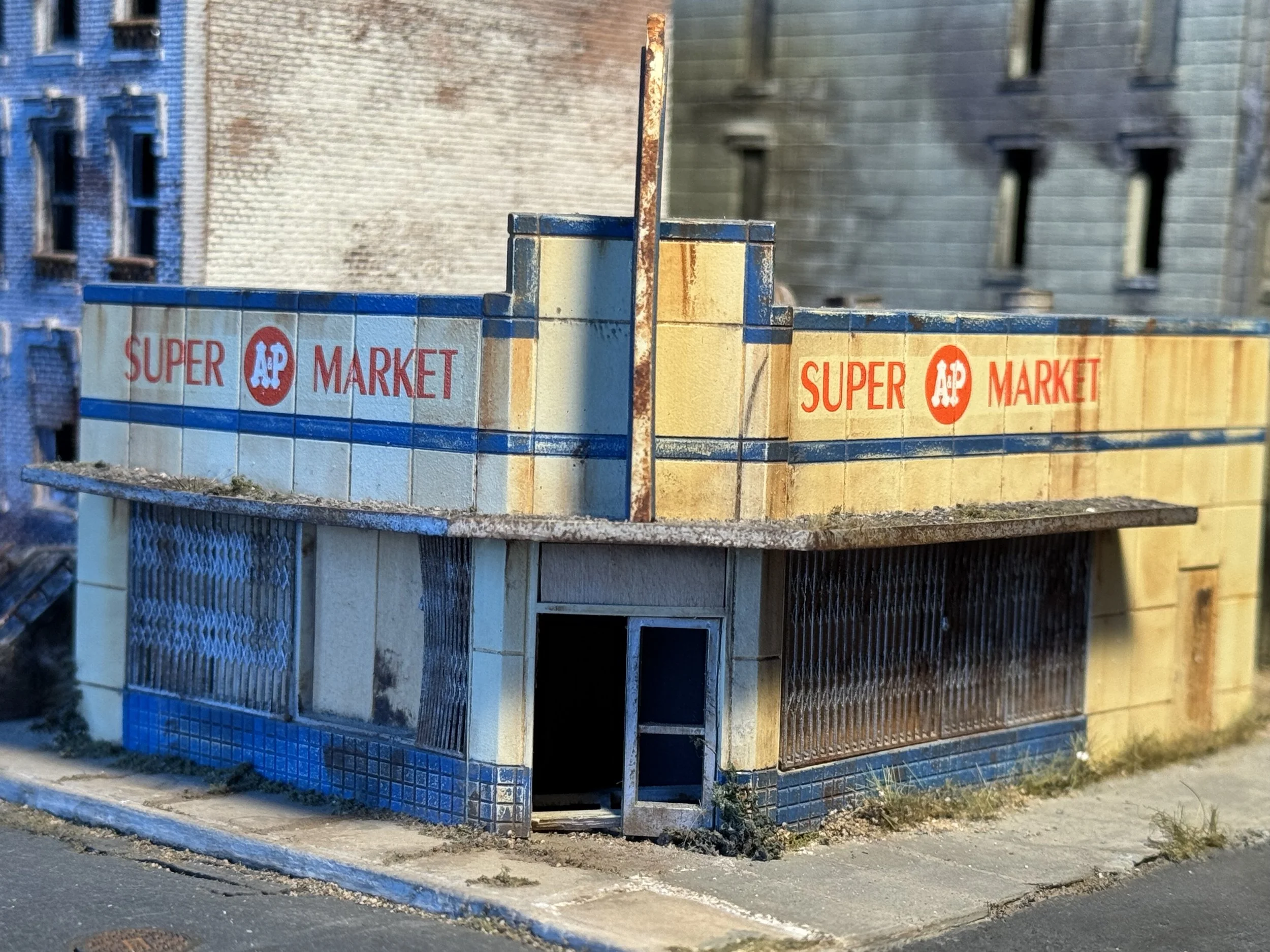 A miniature model of a building with a sign that reads 'Super AP Market' on its front. The model appears weathered and rusty, and the background shows taller, more detailed buildings.