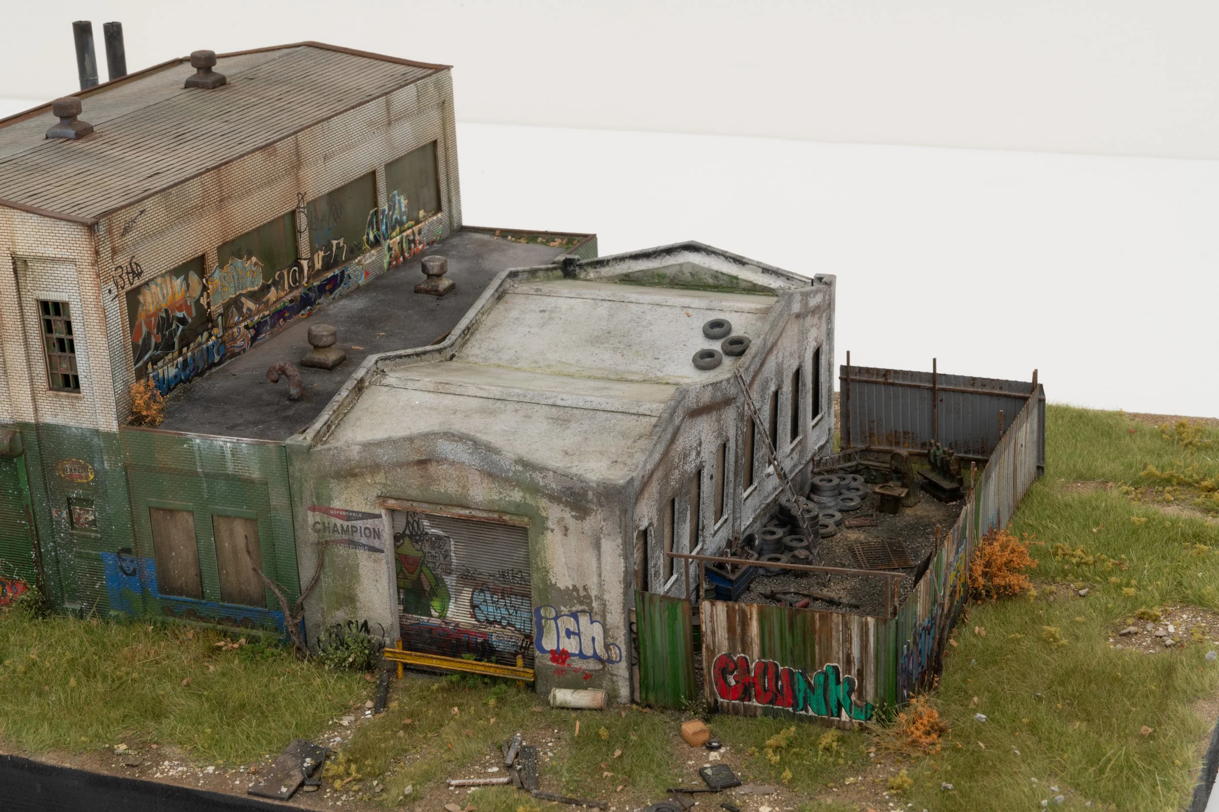 Model of an abandoned industrial building with graffiti on the walls, tires on the roof, and a rusty fence surrounding it, set in a grassy outdoor area.