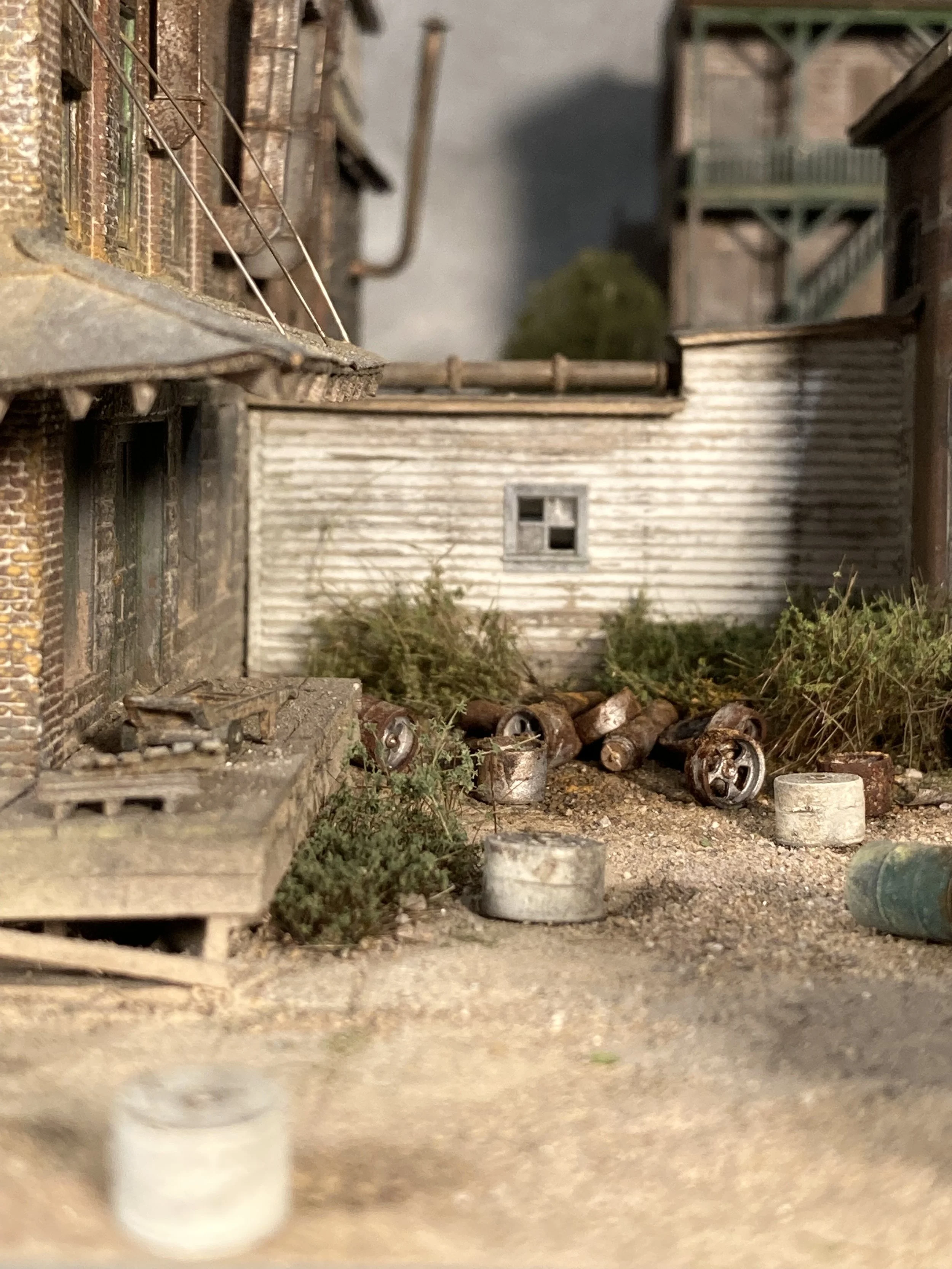 A miniature model of an urban alleyway with buildings, plants, scattered cans and buckets, and debris on the ground.
