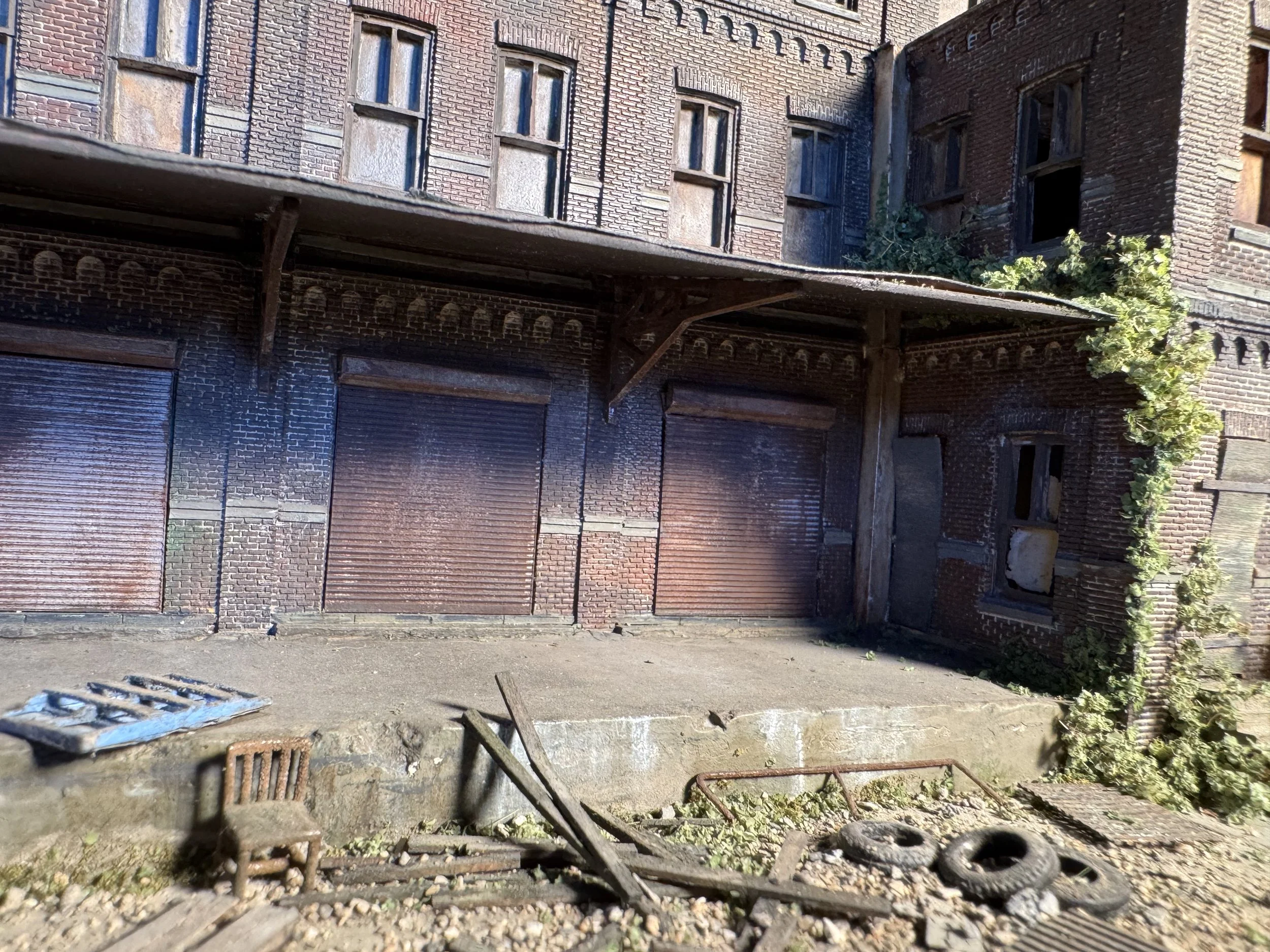 An old brick building with boarded-up windows and a small overhanging roof. The foreground shows some debris and ladders, with concrete ground and some green plants growing next to the building.