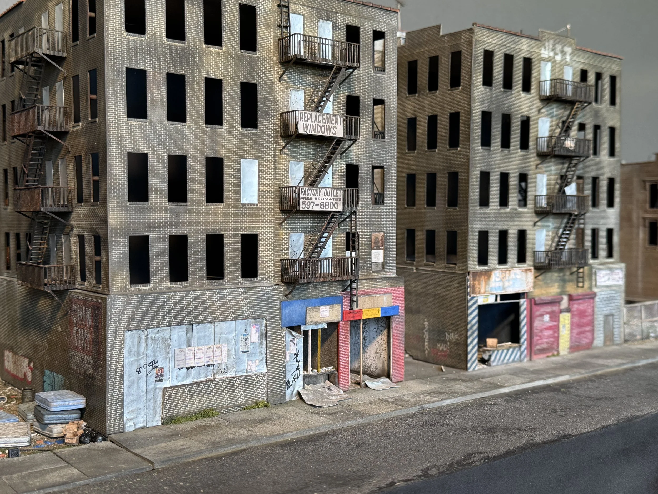 A model of a multi-story brick building with boarded-up windows, fire escape stairs, and street-level storefronts, some of which are boarded up or graffiti-covered.