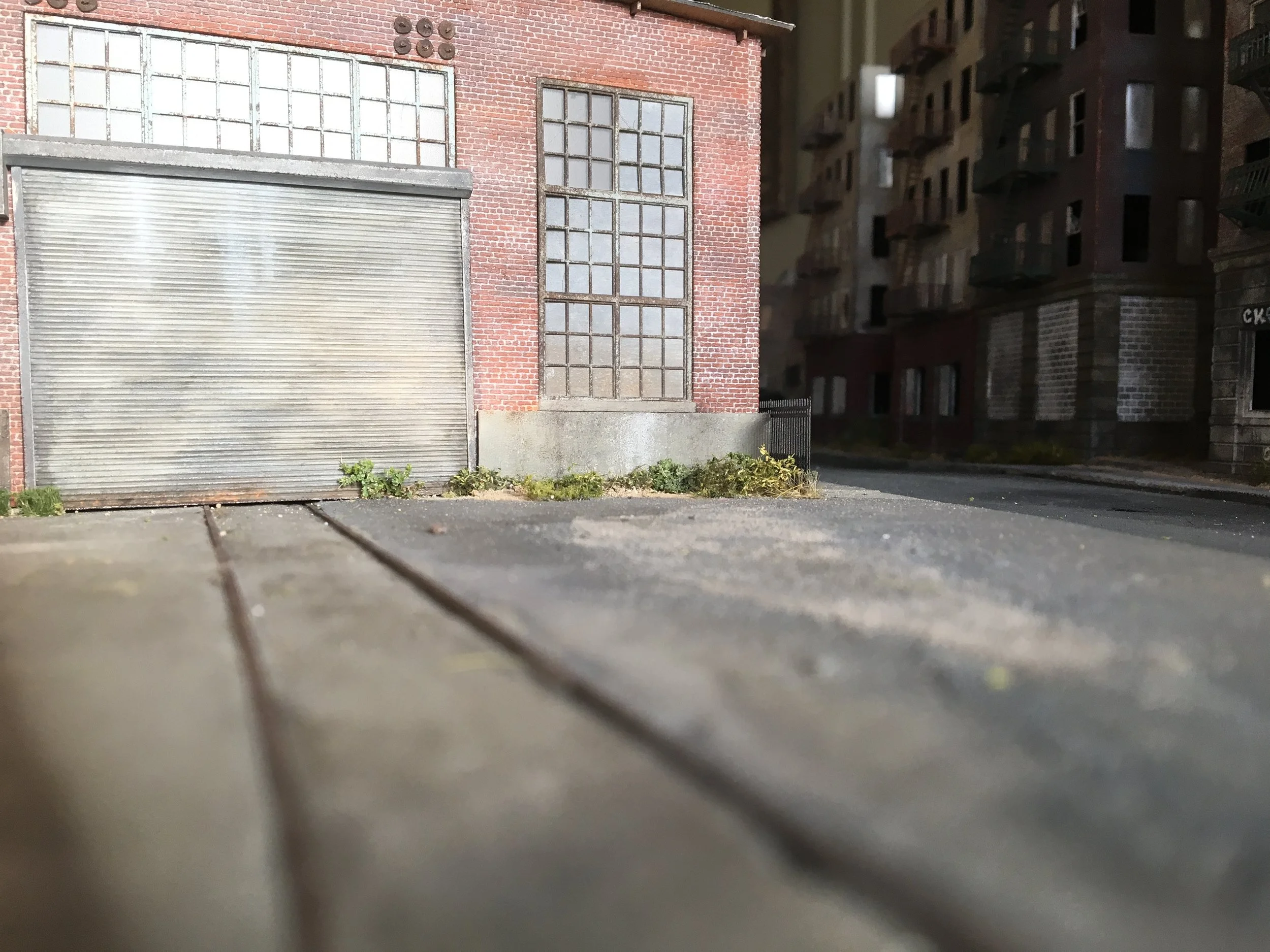 A close-up view of the ground and lower part of a brick building with large windows and a metal roller shutter door, with some plants and weeds growing along the base of the building.