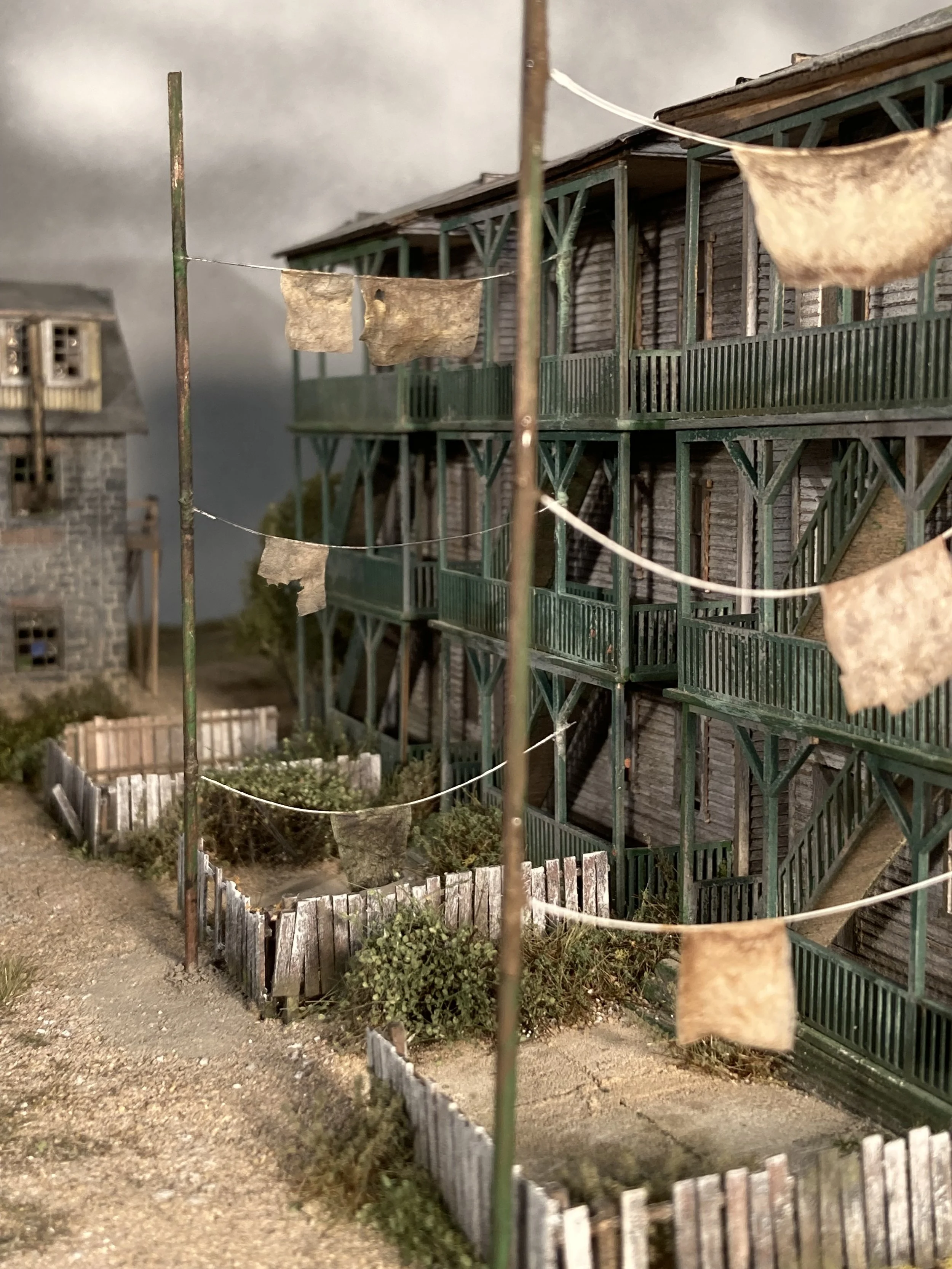 Model of a weathered two-story wooden house with a large porch, surrounded by a small garden enclosed by a rustic wooden fence. Dry clothes hang on a line stretched between two poles in front of the house.