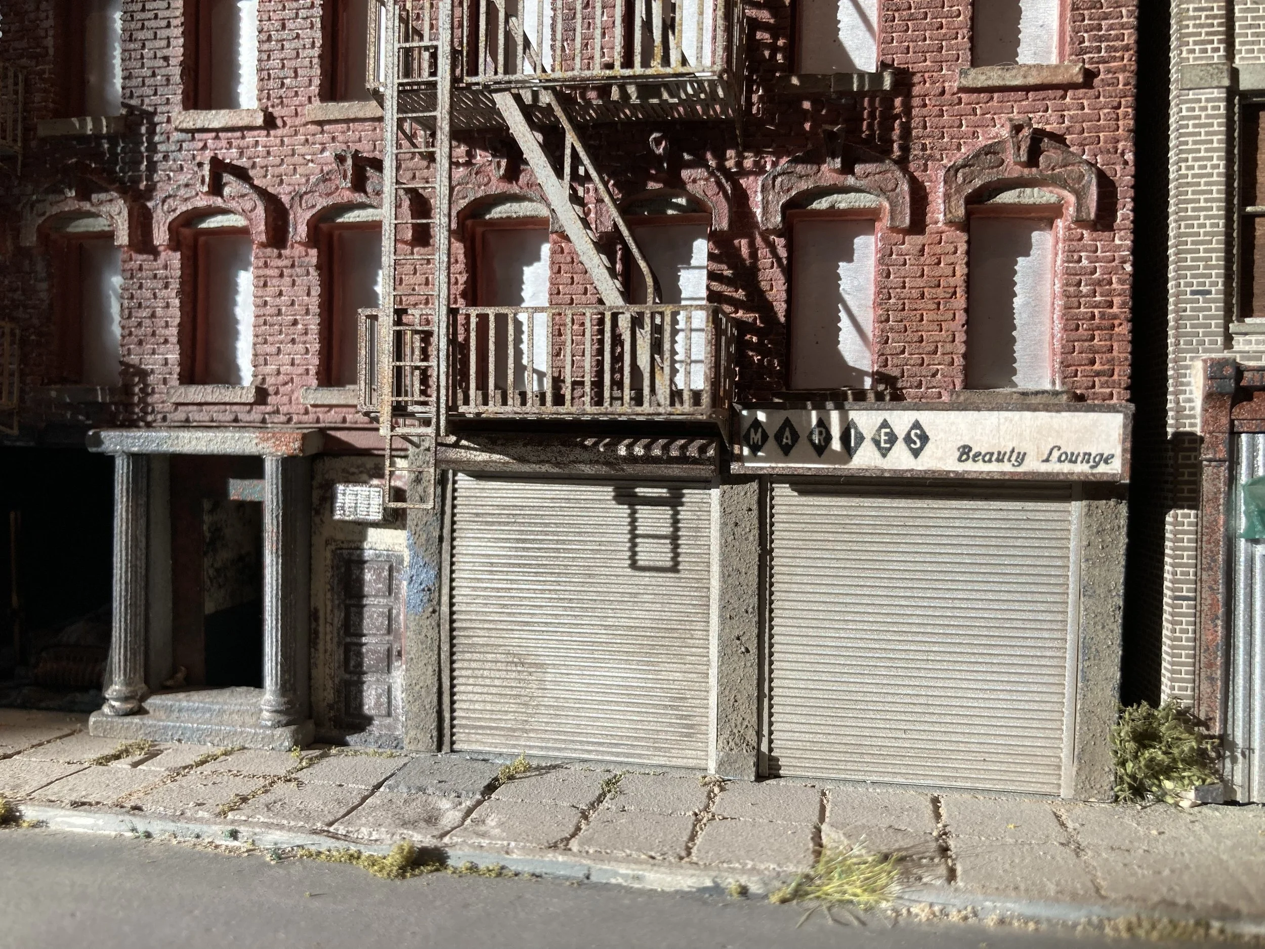 Front view of a brick building with a fire escape staircase and a sign for a beauty lounge, with a closed metal roll-up door and a small plant at the sidewalk.
