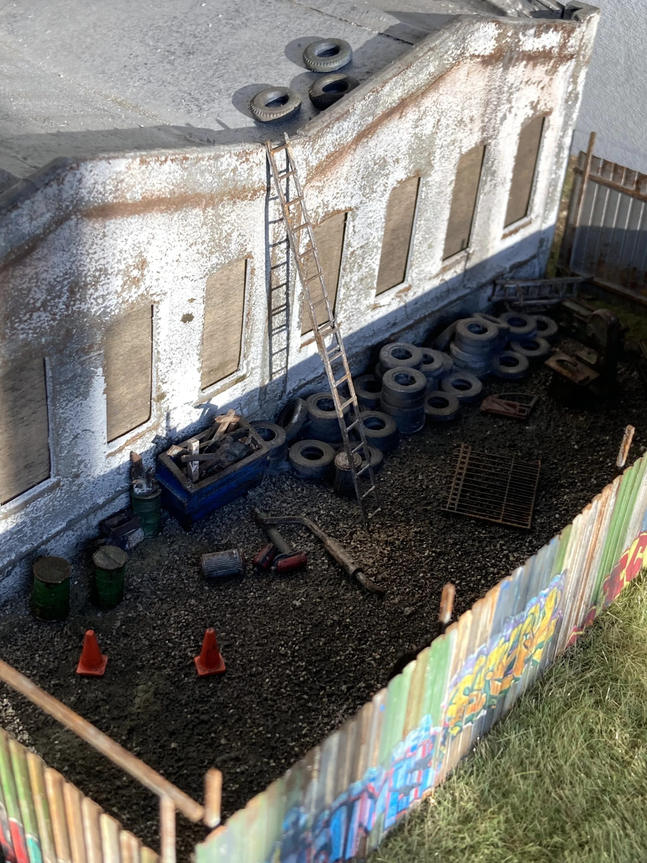 Miniature model of a building with a weathered white exterior, small ladder, and scattered tiny tires and tools inside a colorful fence on a ground covered with dark soil.