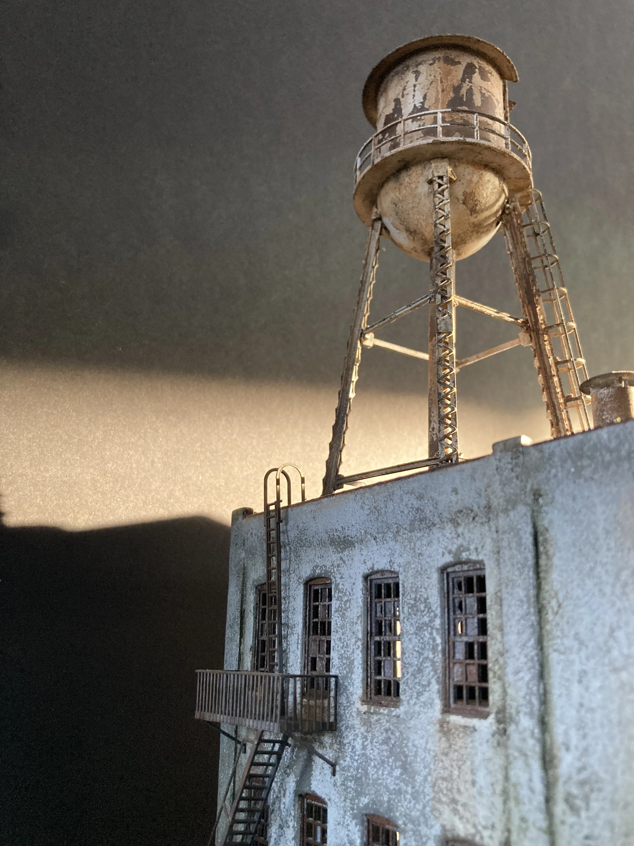 Model of an old industrial building with a water tower on top, featuring rusted metal and weathered white walls, situated under a cloudy evening sky.