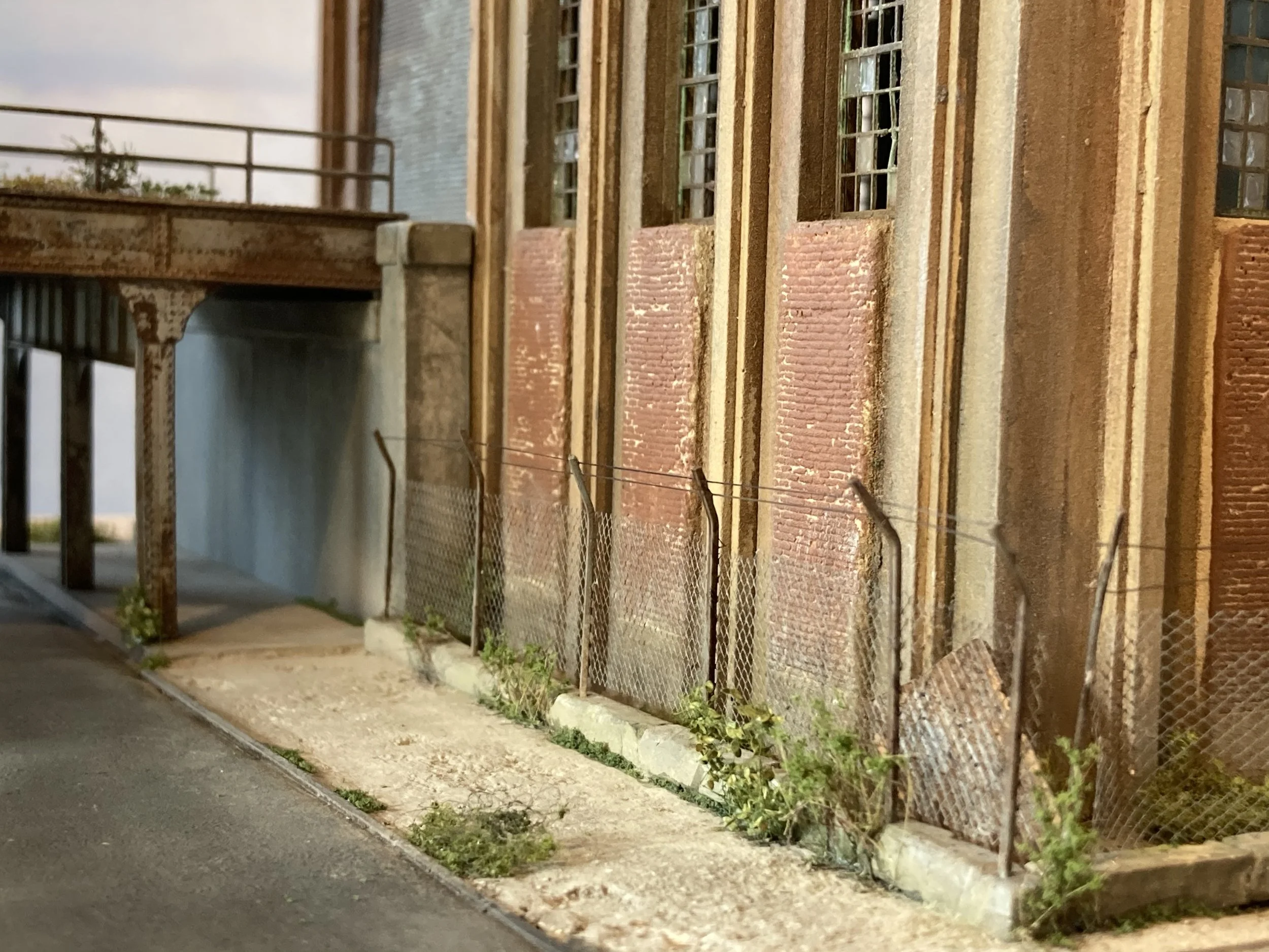 A rusty chain-link fence runs alongside a sidewalk next to a brick and stucco building with multiple windows. The sidewalk has some small plants growing along its edge.