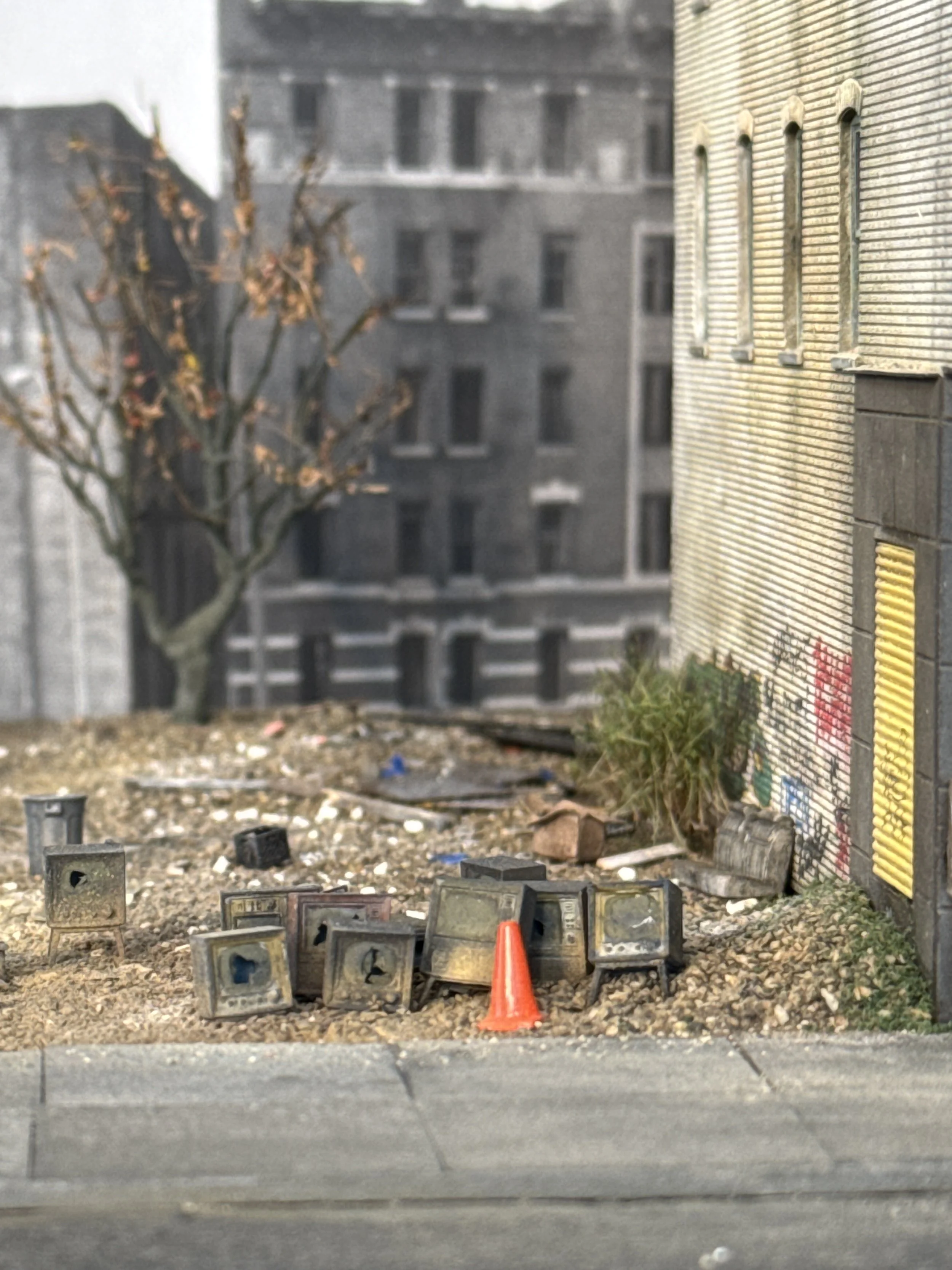 Sidewalk construction site with debris, small cement blocks, an orange traffic cone, a building's wall with graffiti, and a leafless tree in the background.