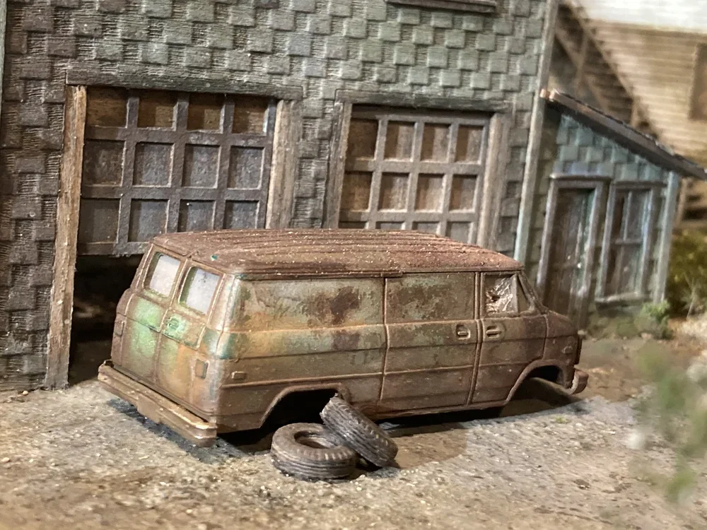 A miniature, rusted toy van parked outside an old, weathered garage with broken tires and worn building exterior.