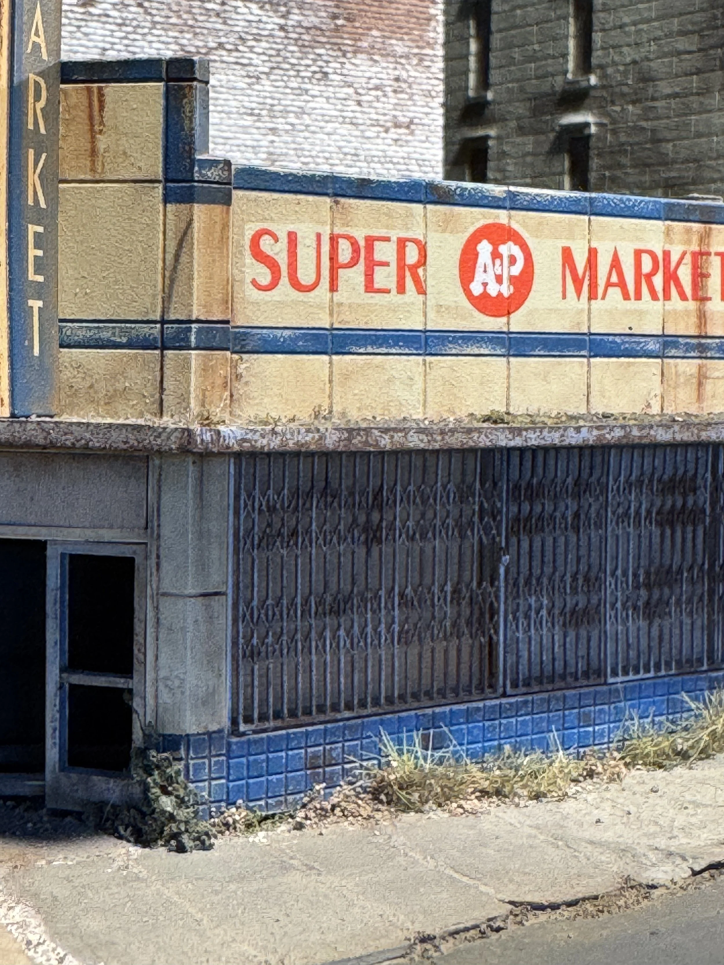 A wall made of beige tiles and dark blue tiles with the words 'ARKE' written vertically on the left side. The wall features a red and white sign with the words 'SUPER MARKET' and a logo in the middle containing the letters 'AP'. In front of the wall,