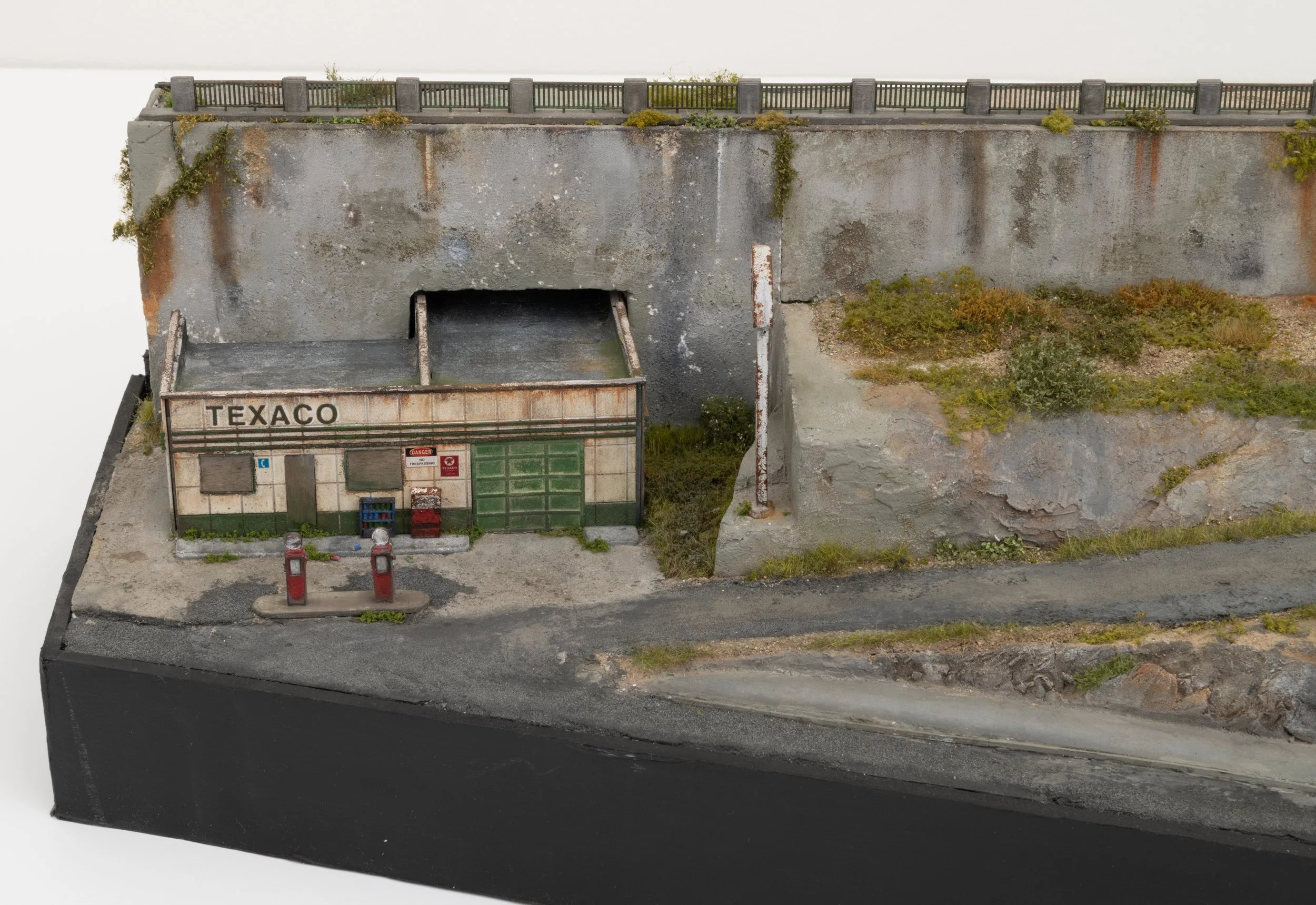 Model of an abandoned, weathered concrete wall with graffiti and moss, featuring a rusted Texaco gas station structure, trash cans, and overgrown vegetation.