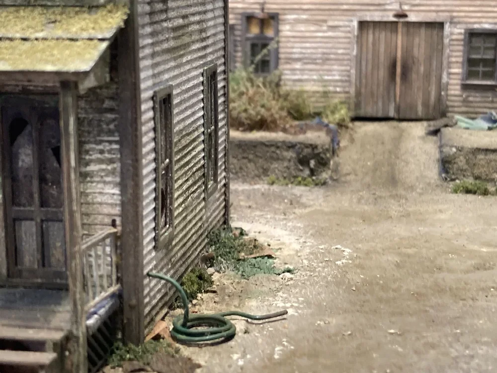Close-up of a weathered miniature model of a rustic wooden house with a garden hose on the ground outside, showing a muddy area and small plants.