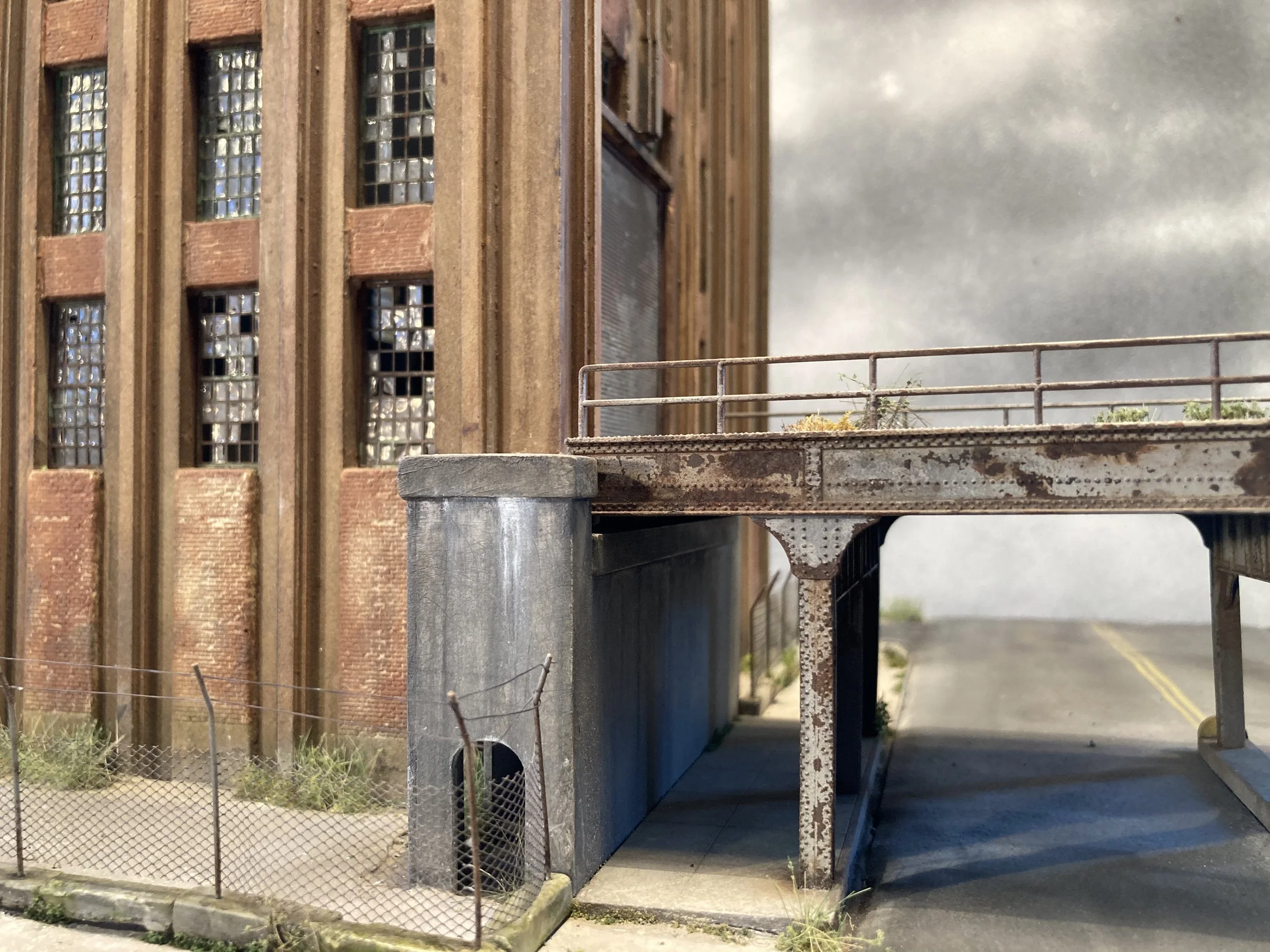 A model of an old industrial building with brick and concrete walls, glass block windows, and an outdoor terrace with a metal railing, located on a street with a sidewalk and a chain-link fence.