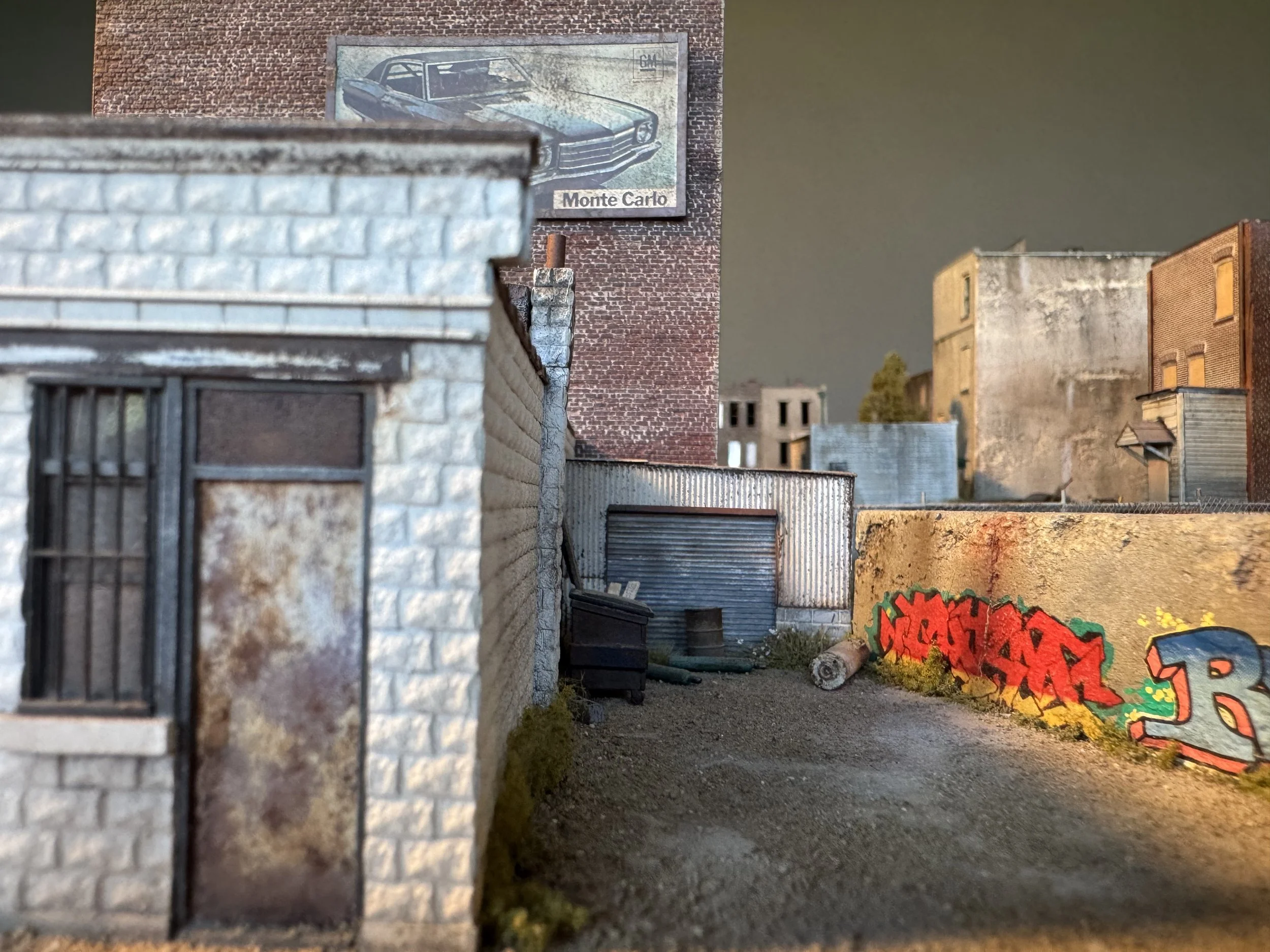 A detailed miniature model of an urban alleyway scene with small buildings, graffiti on a wall, and a backdrop billboard of a classic car labeled 'Monte Carlo'.