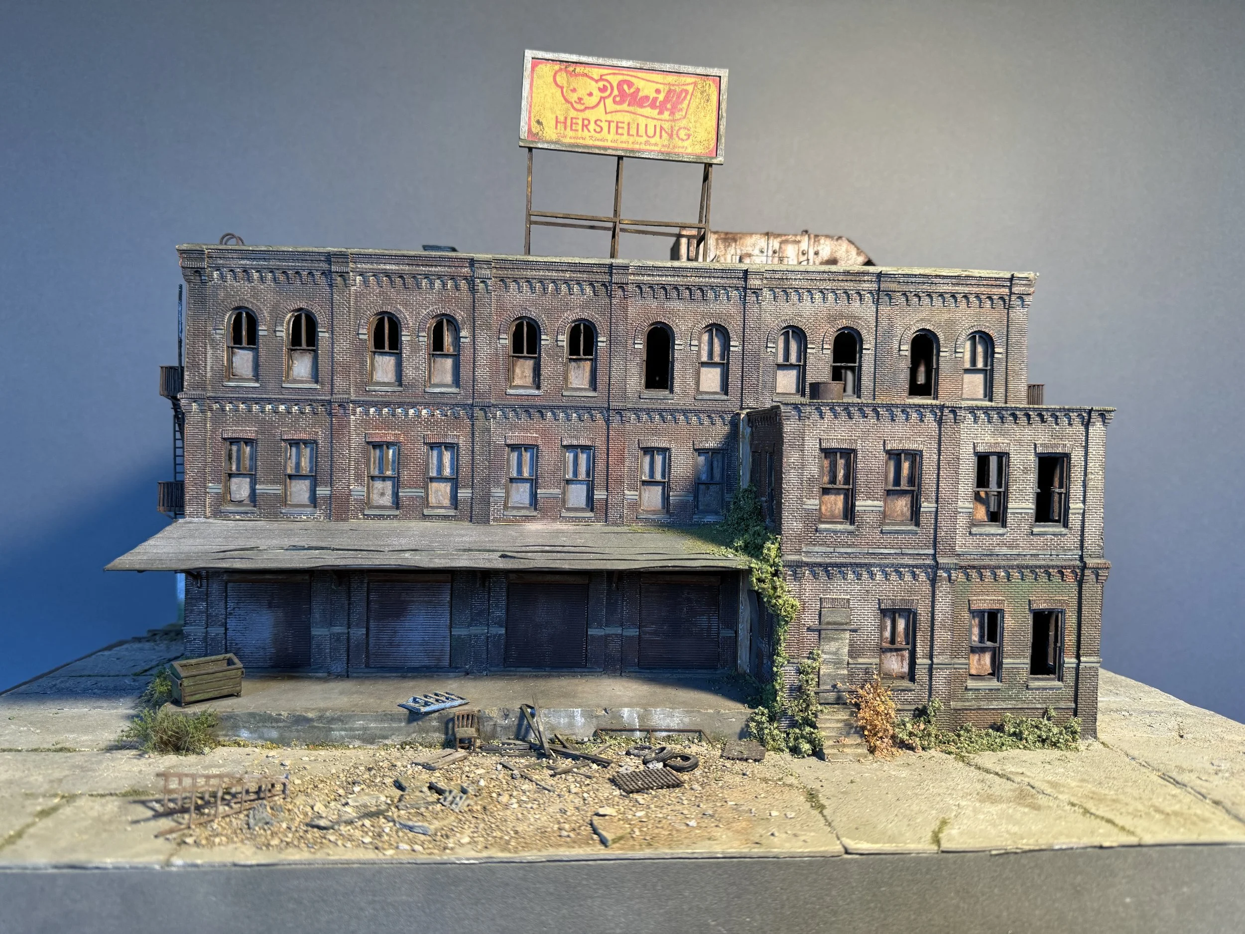 A miniature model of an old brick building with a billboard on top, set on a textured base with small plants, debris, and a crate in front, against a plain dark background.