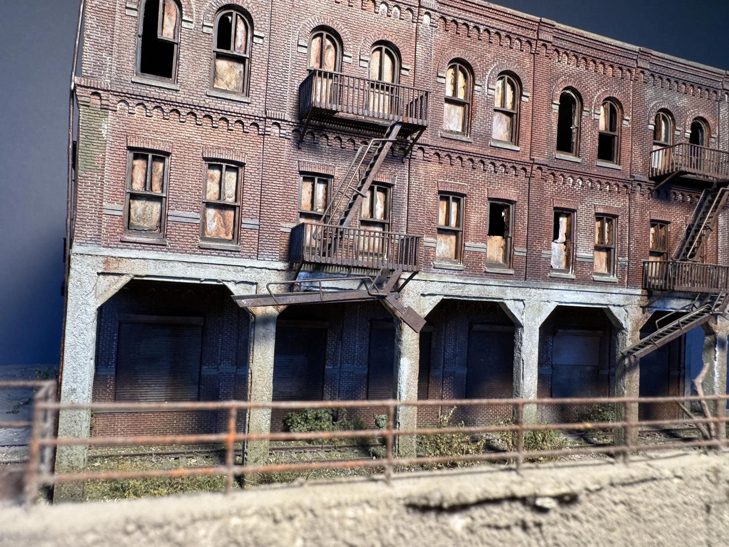 A detailed model or miniature of an old brick building viewed through a wire fence. The building has multiple windows, some open or broken, fire escapes on the exterior, and an arched entrance at the bottom.