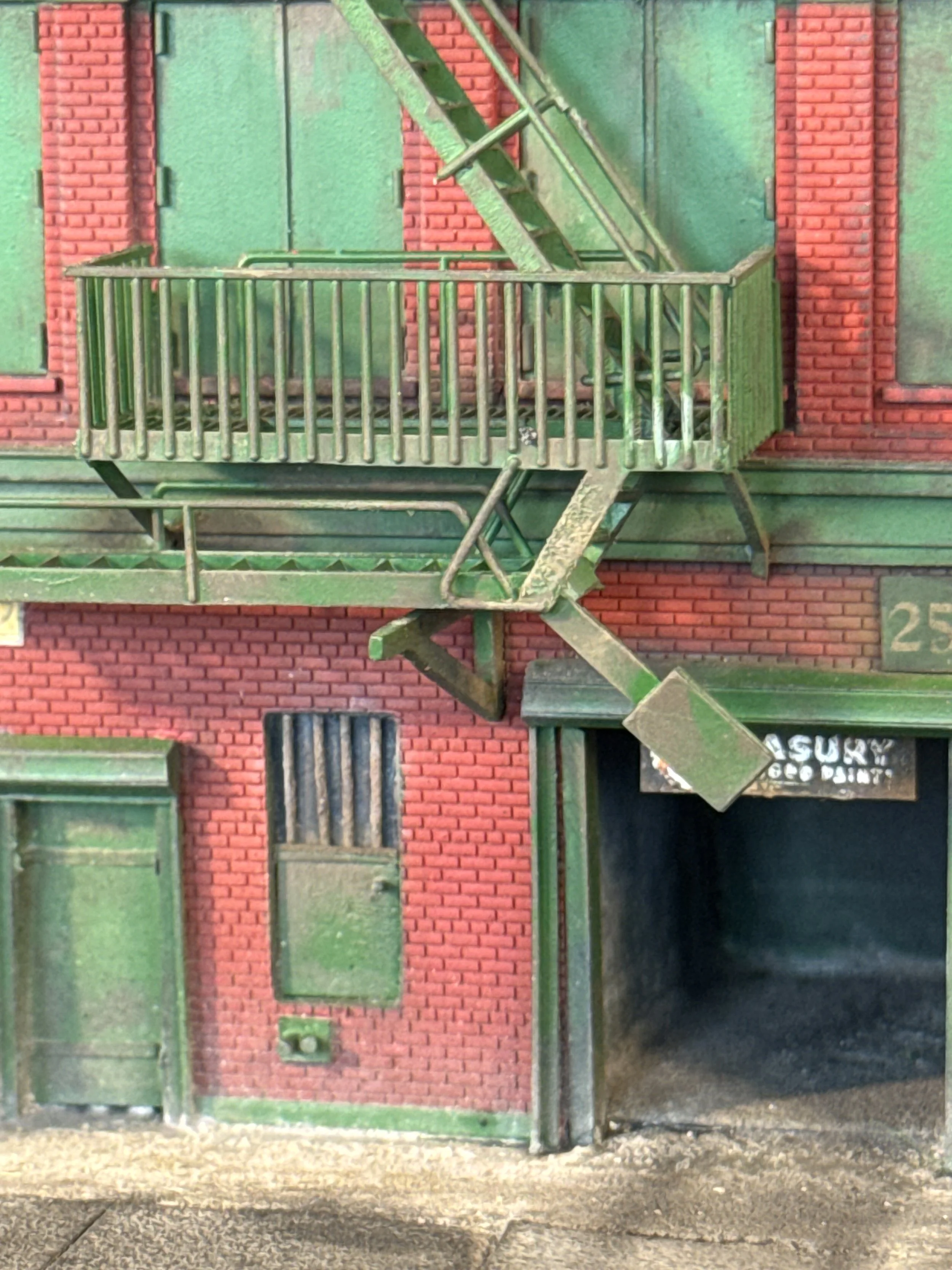 The image shows a building façade with red brick walls and green fire escape stairs attached. There is a door on the upper level, and windows on the lower level, one with blinds. The lower part of the image includes a black garage door with partially