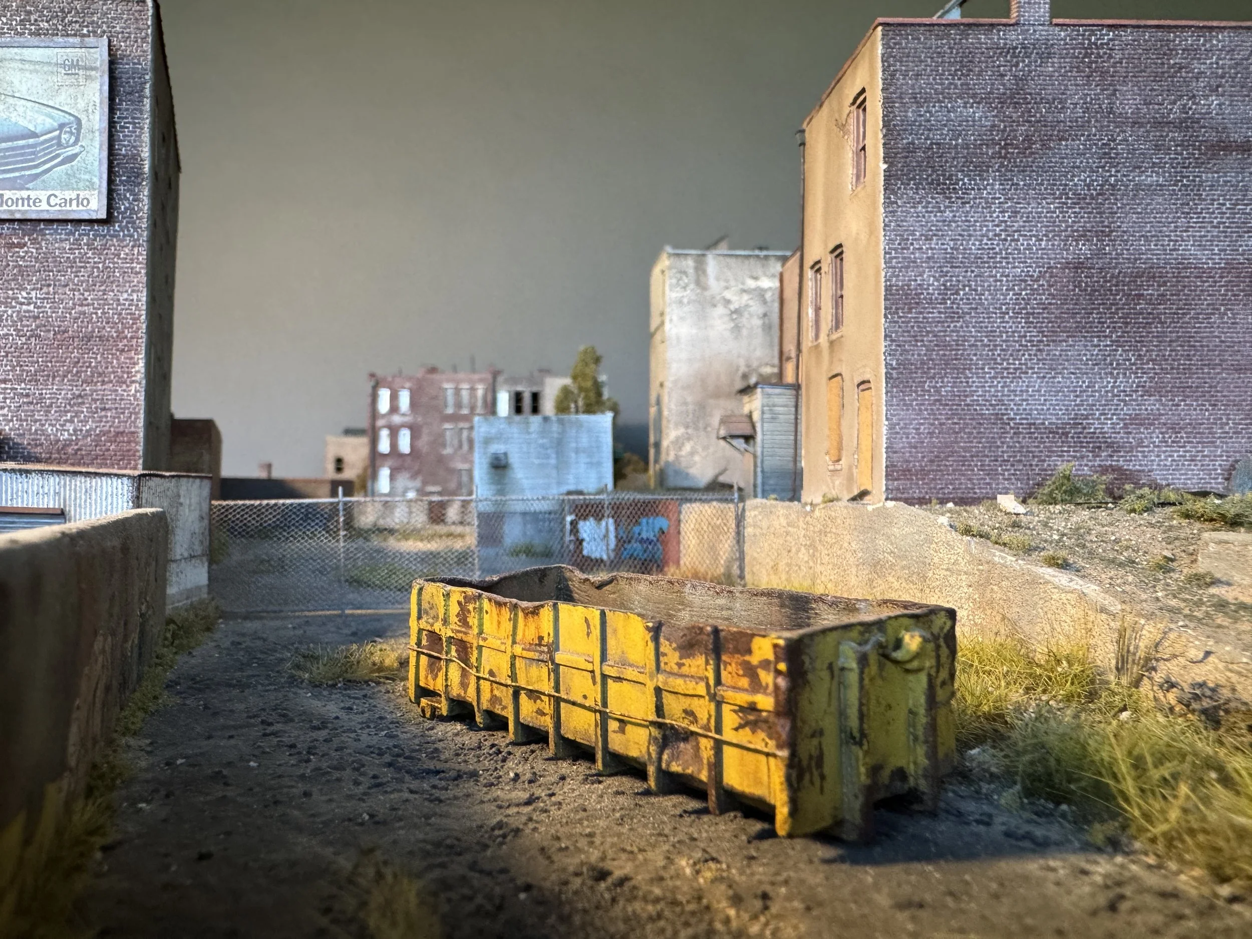 Miniature model of an urban alleyway scene with buildings, fences, and a yellow barrier in the foreground.