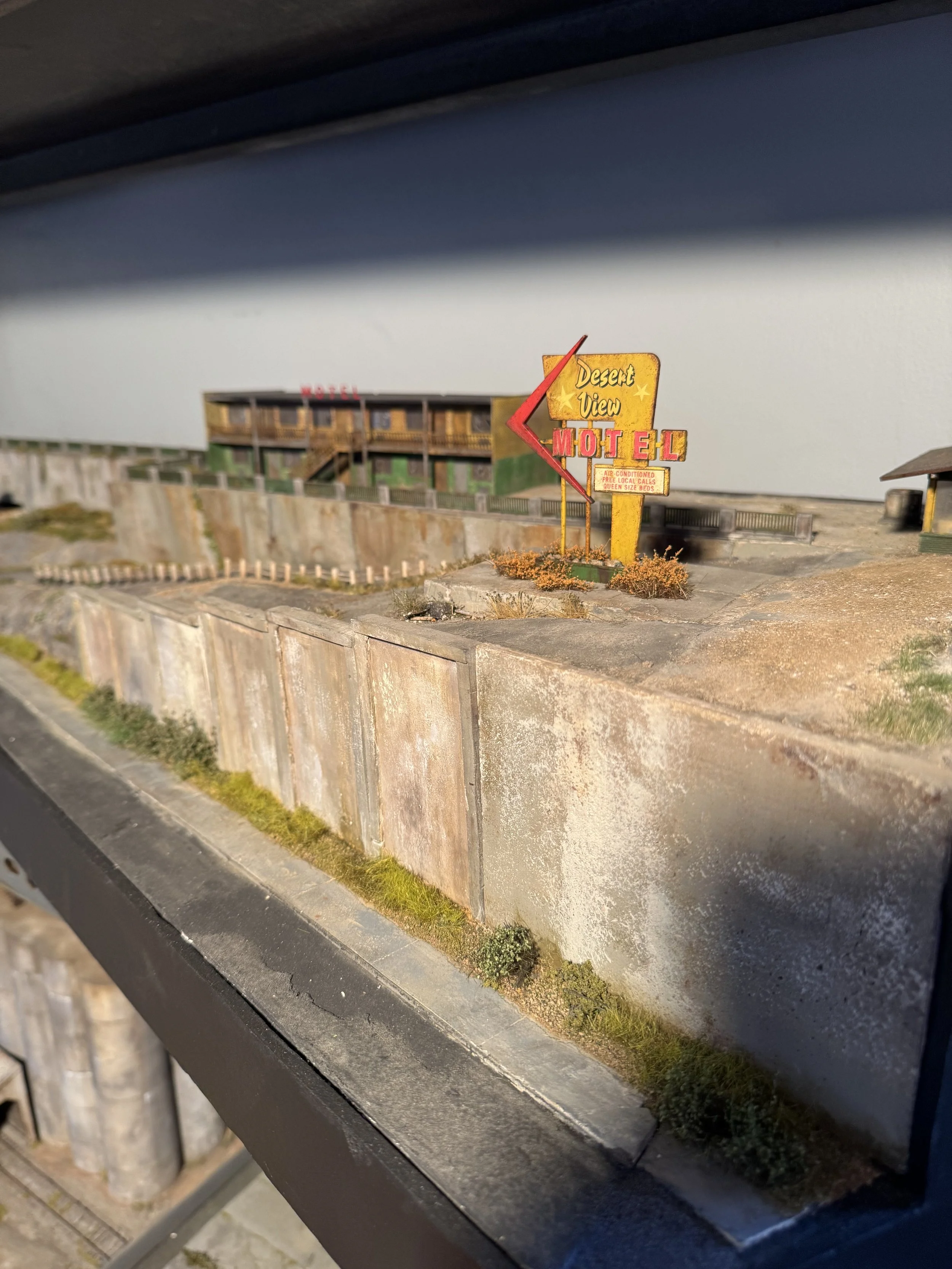Miniature model of a desert motel with a yellow and orange sign and a two-story wooden building in the background, situated near a concrete wall and grassy area.