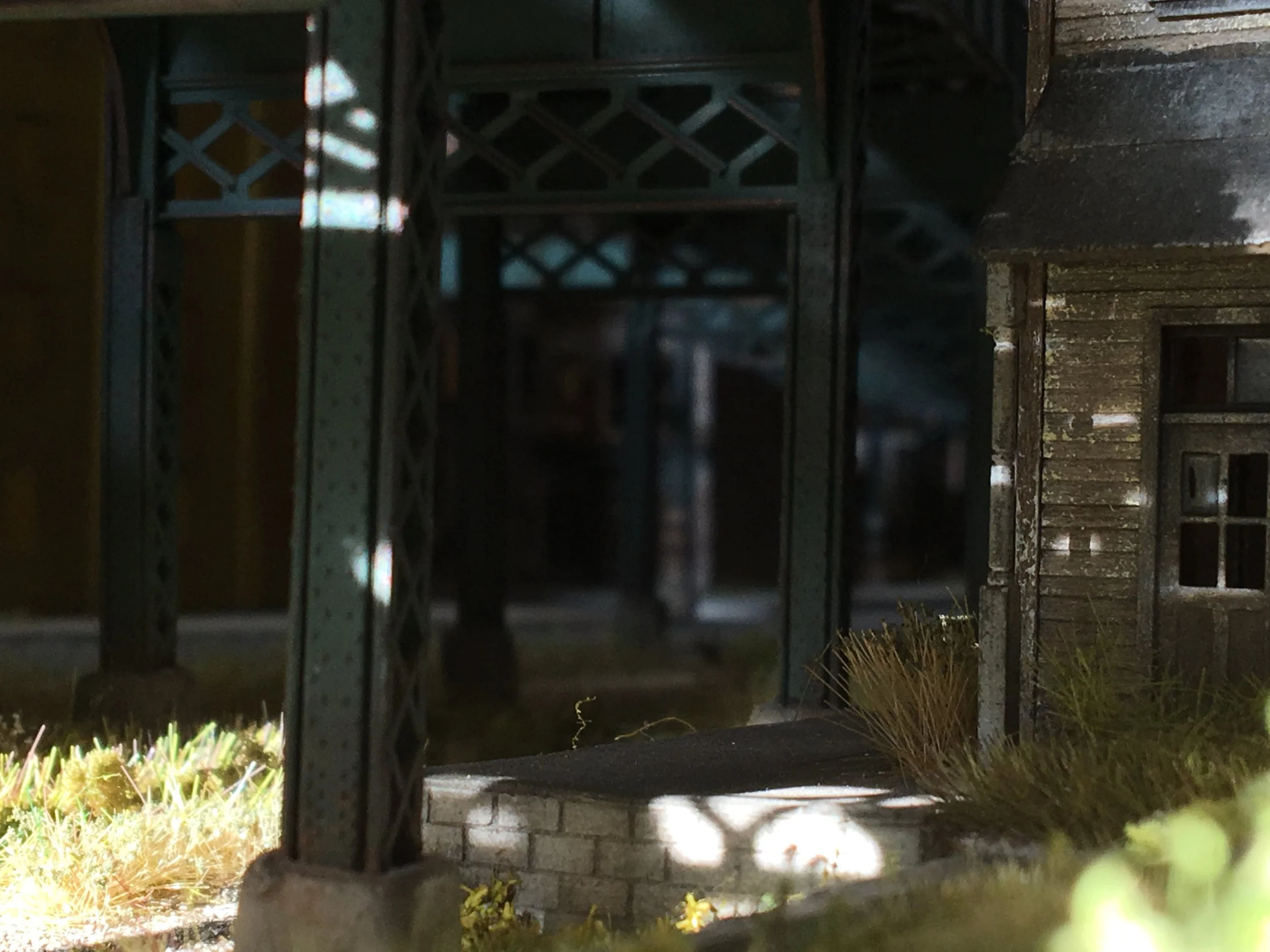 A dark night scene showing a lit street with a house, some plants, and a metal structure, possibly part of a fence or gate, with shadows cast on the ground.
