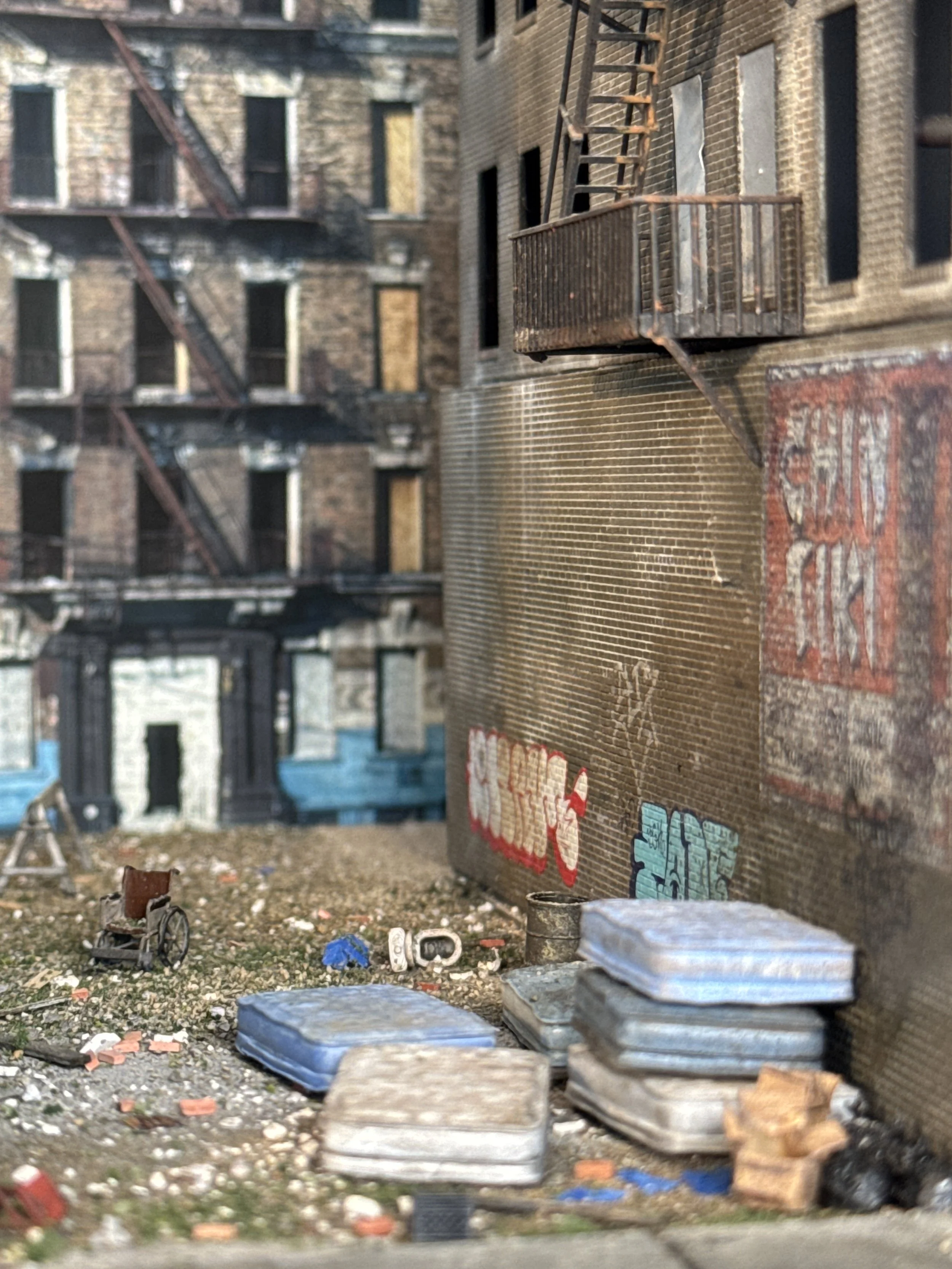 A close-up view of a miniature model of an urban alleyway scene with detailed brick buildings, fire escapes, graffiti on the wall, and various small objects and debris on the ground.