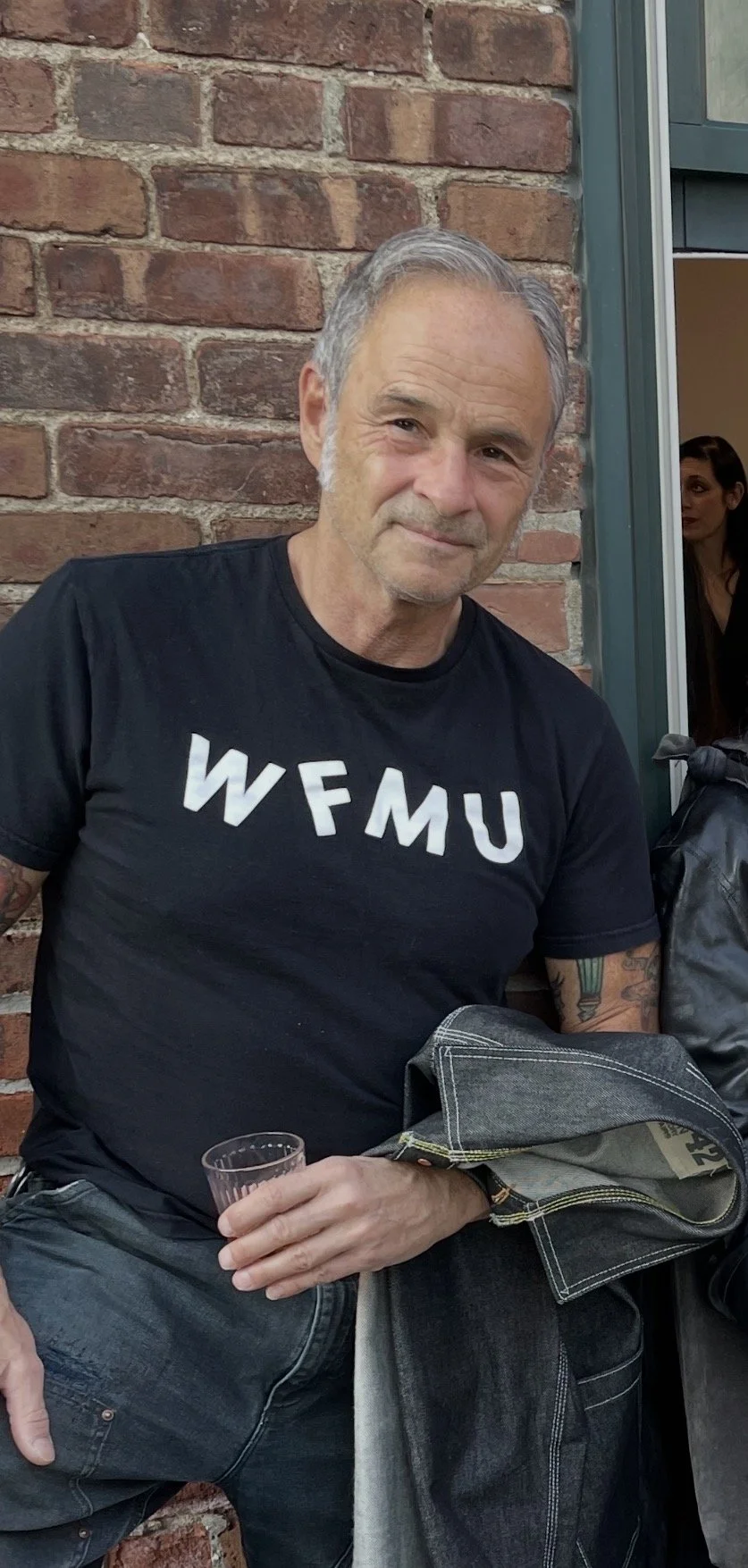 A man with gray hair and a beard wearing a black T-shirt with white text reading 'WFMU', sitting outdoors against a brick wall, holding a clear glass. There is a woman partially visible in the background near a window.