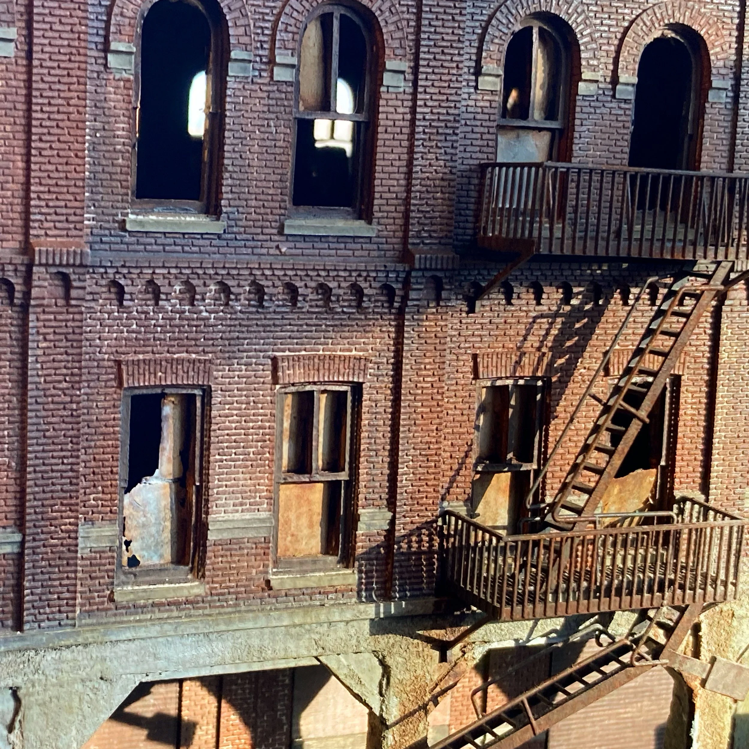 A brick building with multiple arched windows, some of which are boarded up or missing glass. There is a fire escape attached to the right side of the building, casting shadows on the brick wall. The building appears to be old and partially dilapidat