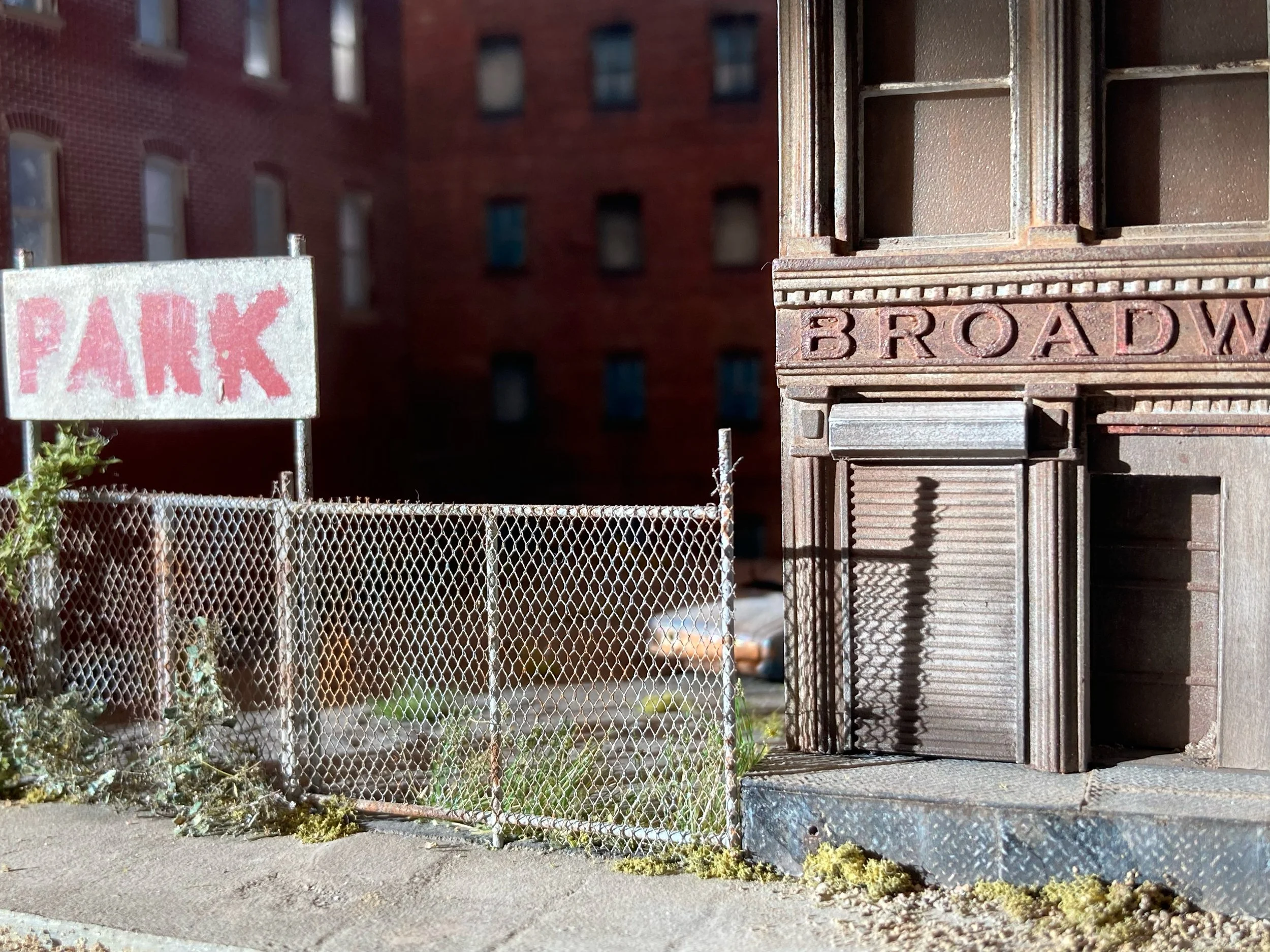 A metal fence with a patched hole, a 'PARK' sign, and a rusted fire exit door on a city sidewalk with a brick apartment building in the background.