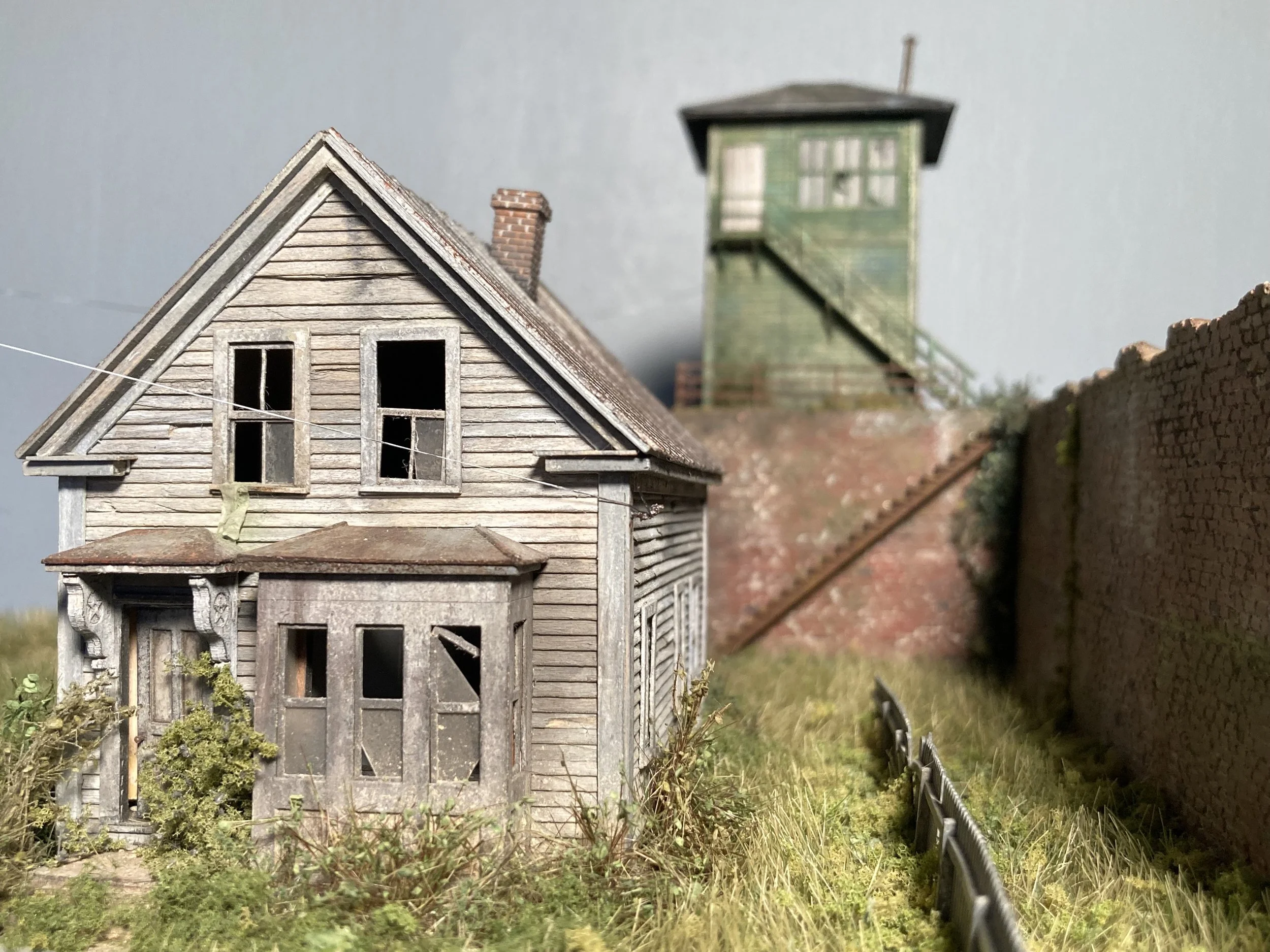 Close-up of a weathered miniature wooden house with a gabled roof and multiple windows, with a blurred green watchtower in the background.