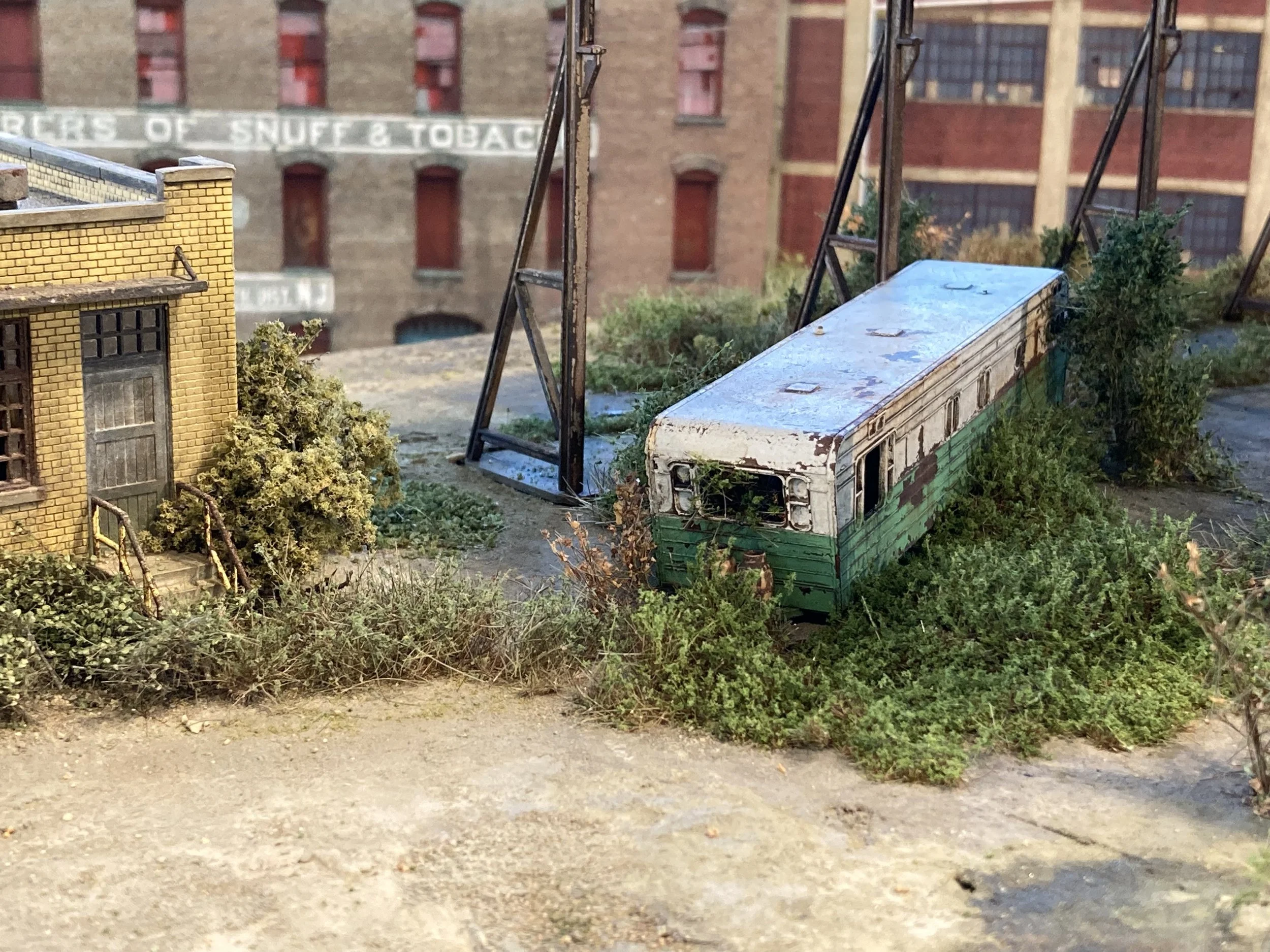 Miniature model of an outdoor urban scene with a yellow brick building, greenery, a bus, and metal structures, set against a background of a larger brick building with windows and a sign.