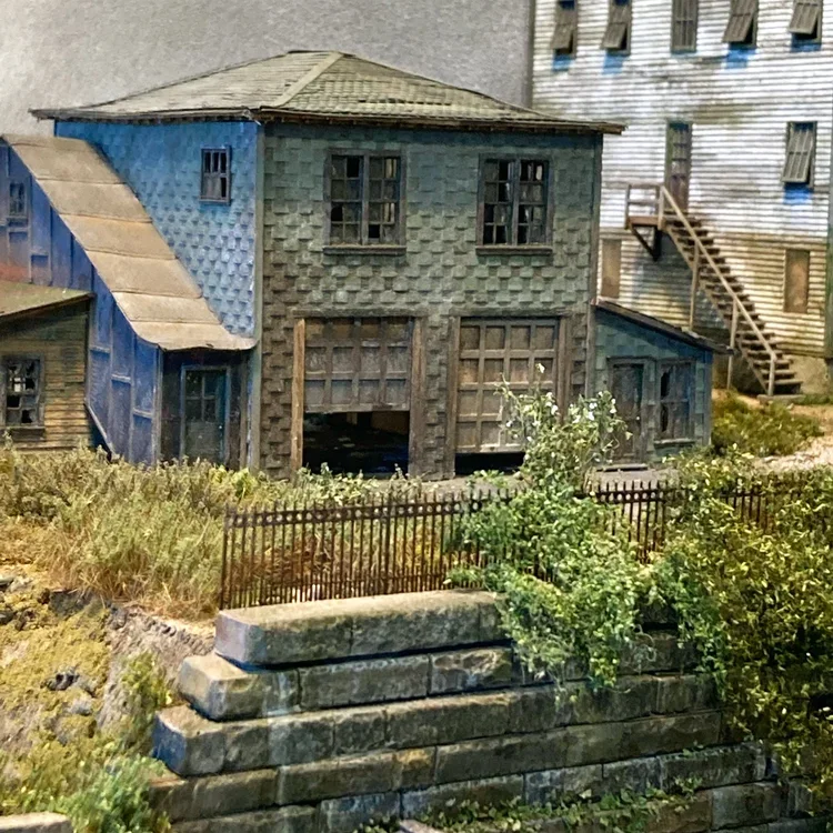 Miniature model of a two-story house with a brown shingled roof, blue and green exterior walls, and a wooden staircase on the right side. There is a small fence and some greenery in the foreground.