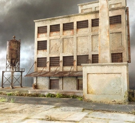 A weathered, abandoned four-story industrial building made of concrete with several barred windows and a rusted awning. To the left, a tall, rusted water tower stands with a cloudy, ominous sky in the background.