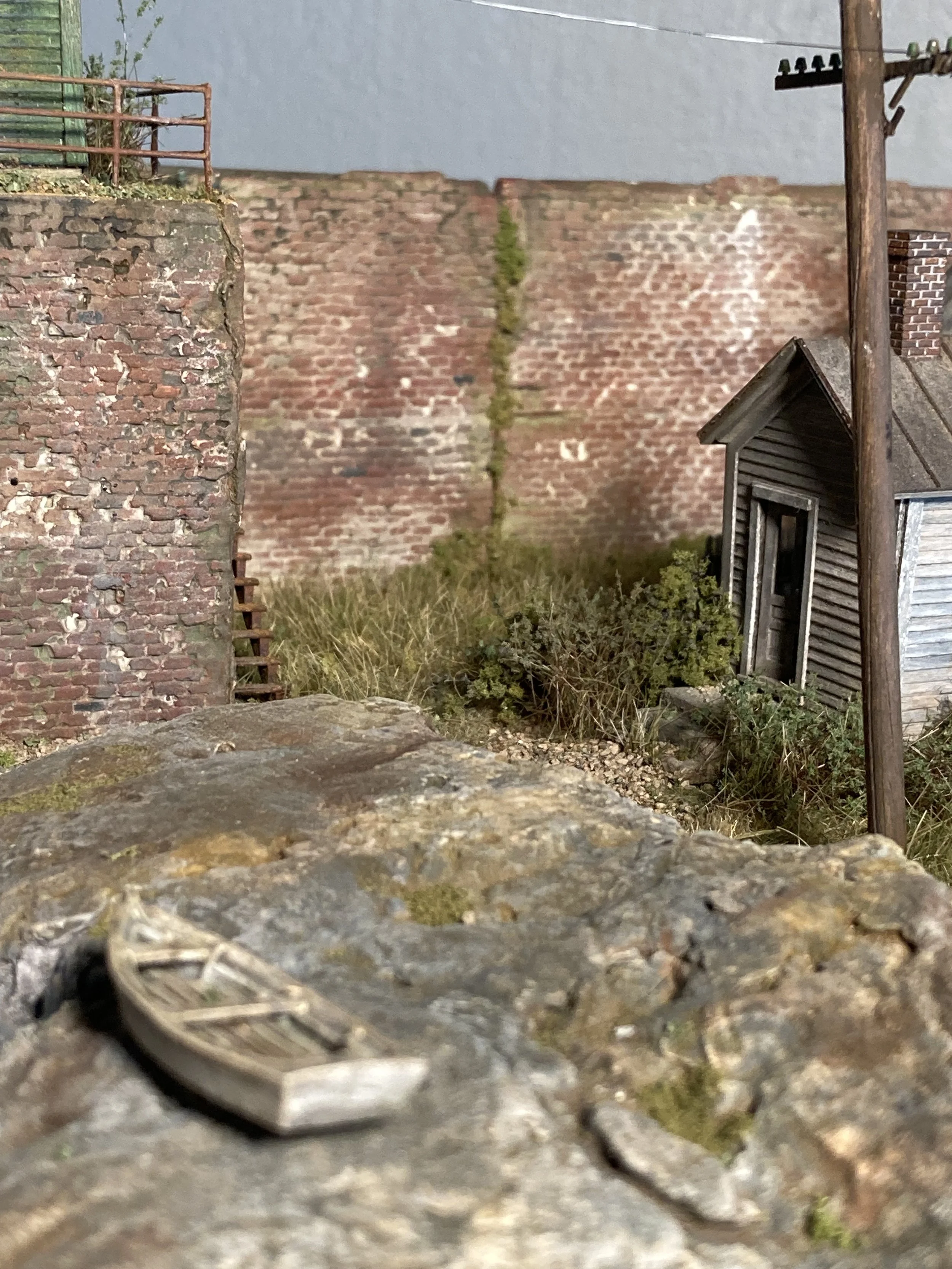 Close-up of a worn coin lying on a rough outdoor surface with gravel and moss in the foreground. In the background, there are brick walls, a wooden house, utility poles, and a cloudy sky.